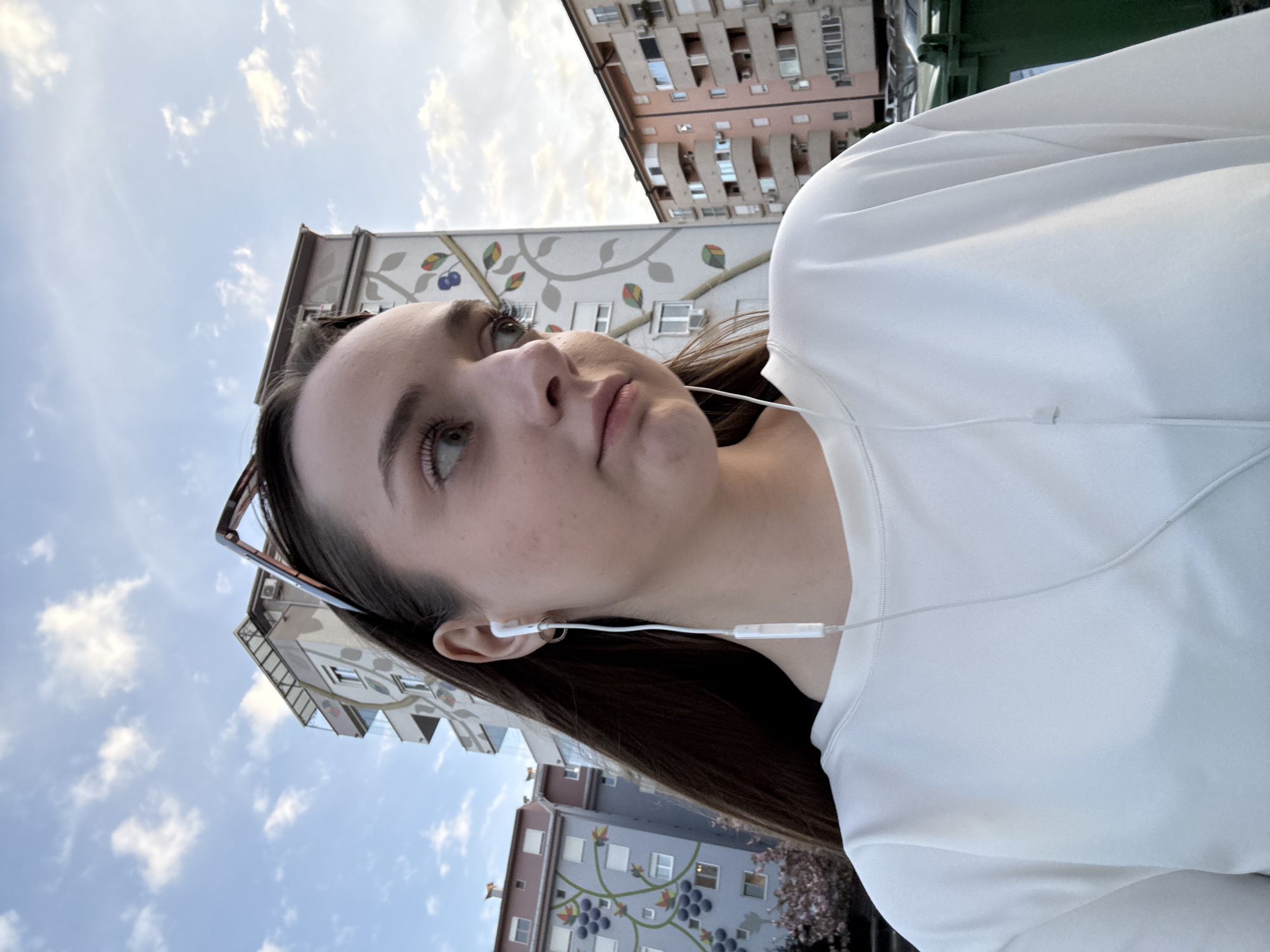 A medium shot, taken from a low angle, depicts a young woman wearing a white top and white earphones, looking up and to the left. She is standing outside in an urban area. In the background, there are several buildings. One building, located behind her, is decorated with murals depicting leaves and fruit. Another building, partially visible at the top, is a light pink color and has several windows and air conditioning units. The sky is visible in the background, with some clouds present. The image was taken in Kragujevac, Serbia.