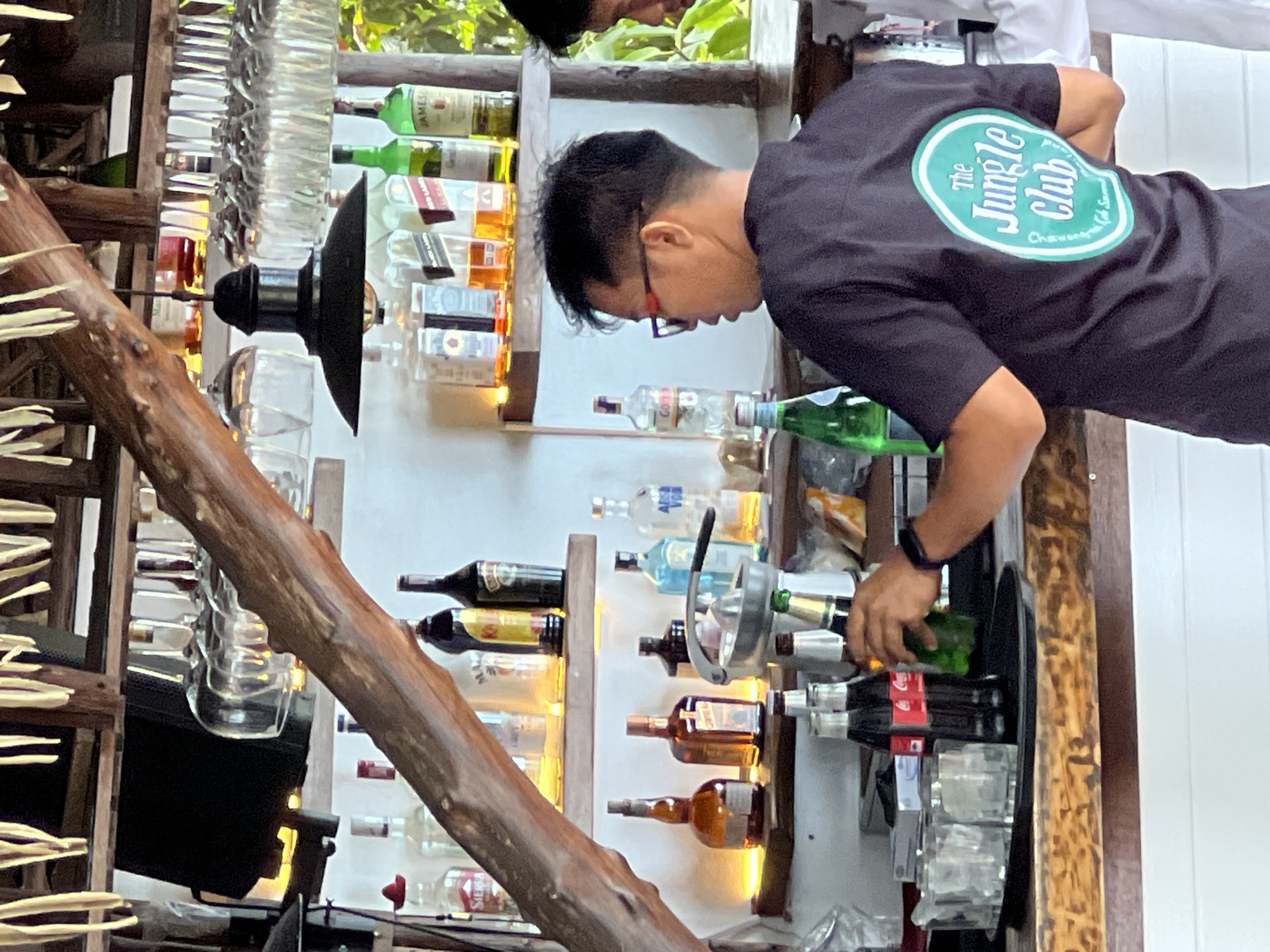 A man wearing a black t-shirt with a green oval logo that says "The Jungle Club" is standing behind a wooden bar in Baan Chaweng Noi, Thailand. He is holding a green bottle and appears to be serving drinks. Behind him, shelves are stocked with various bottles of alcohol, glassware, and a juicer. The bar area is decorated with natural elements, including a large wooden beam and thatched roofing. The overall scene depicts a bar environment with staff preparing drinks for customers.