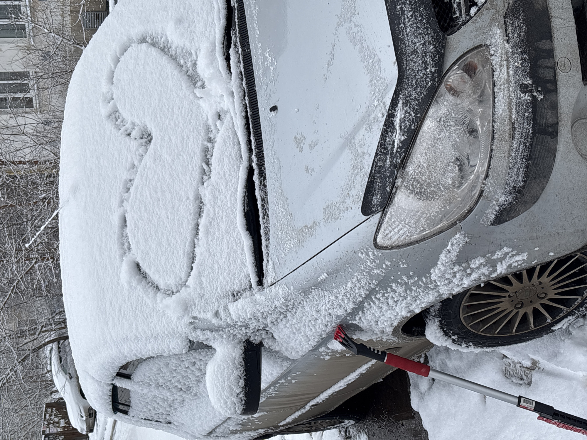 The image shows the front of a snow-covered silver car. Someone has drawn a heart in the snow on the hood. A snow brush with a red handle is present near the front tire. The car appears to be parked outside, with a building and bare trees visible in the background. Based on the appearance of the buildings, it is plausible that the location is Izhevsk, Russia.