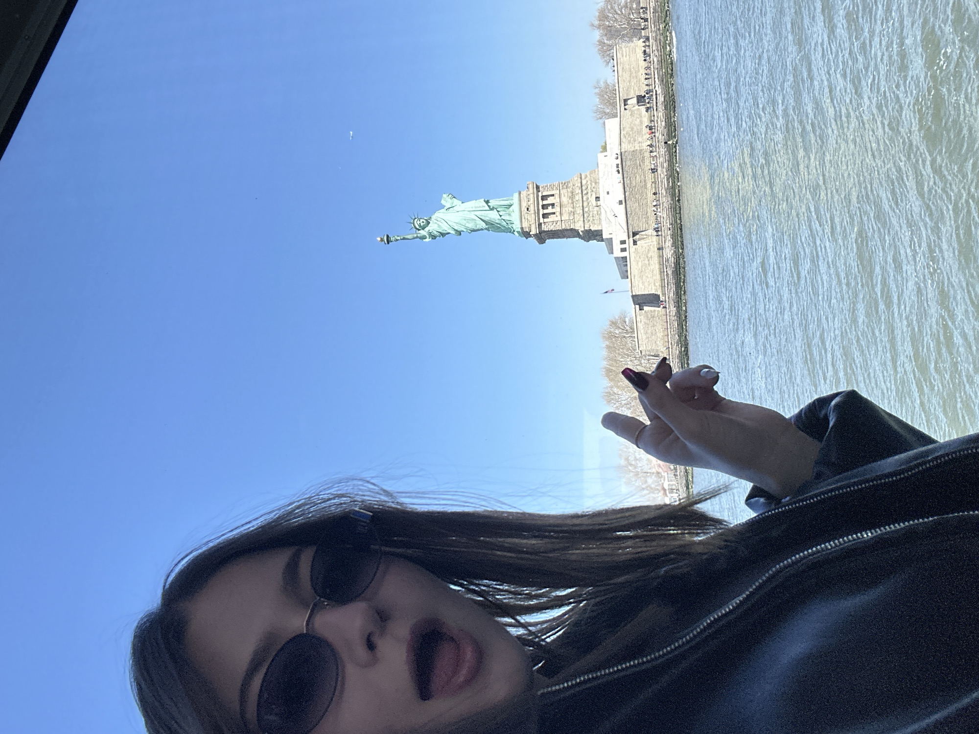 A person wearing sunglasses and a black jacket is taking a selfie with the Statue of Liberty and Liberty Island in the background. The Statue of Liberty stands tall on Liberty Island, with a clear blue sky above. The person has their tongue out and is holding their hand up. The water is visible to the side of the island.