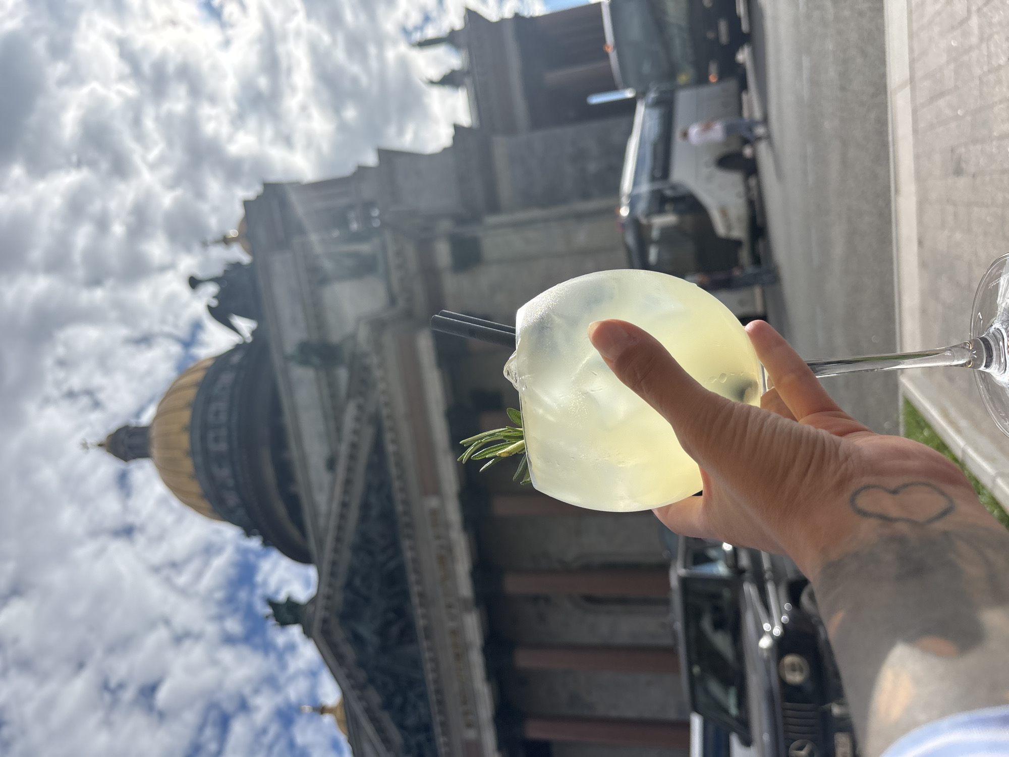 The image shows a hand holding a translucent cocktail with ice and a sprig of rosemary. Two black straws are in the drink. The background is out of focus but appears to be a large, ornate building with a golden dome, possibly a cathedral or a significant historical structure. The sky is cloudy with patches of blue. The lighting suggests it is daytime, likely mid-afternoon due to the bright but slightly diffused sunlight. The hand holding the drink has a visible tattoo of a heart and other shaded designs. The scene seems to be outdoors, possibly a cafe or an area with a view of the architecture. There is no visible text in the image. The overall mood is relaxed and summery, with a focus on enjoying a drink in a scenic location.