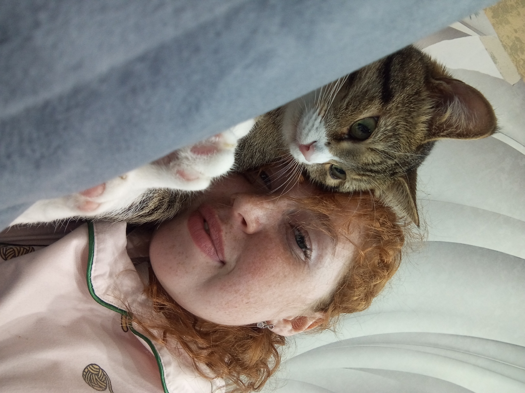 A light-skinned person with freckles and red hair is lying down wearing a light pink pajama top with a yarn ball design. A tabby cat with green eyes and white paws is on top of the person. A gray blanket is partially covering the upper left corner of the image. The background is white with curved lines. The location is not determinable from the visual elements of the image.