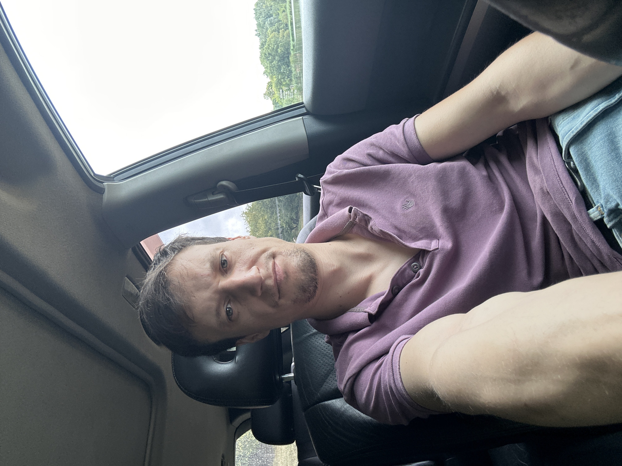 A man is pictured from the chest up, sitting in the passenger seat of a car. He has short brown hair and is wearing a purple polo shirt and blue jeans. The man is looking towards the camera. The interior of the car is visible, including the black leather seats and the sunroof. Outside the car, trees and a body of water are visible. The image appears to have been taken on a cloudy day. The image does not provide specific information about the location in the city of Makiv, Ukraine.