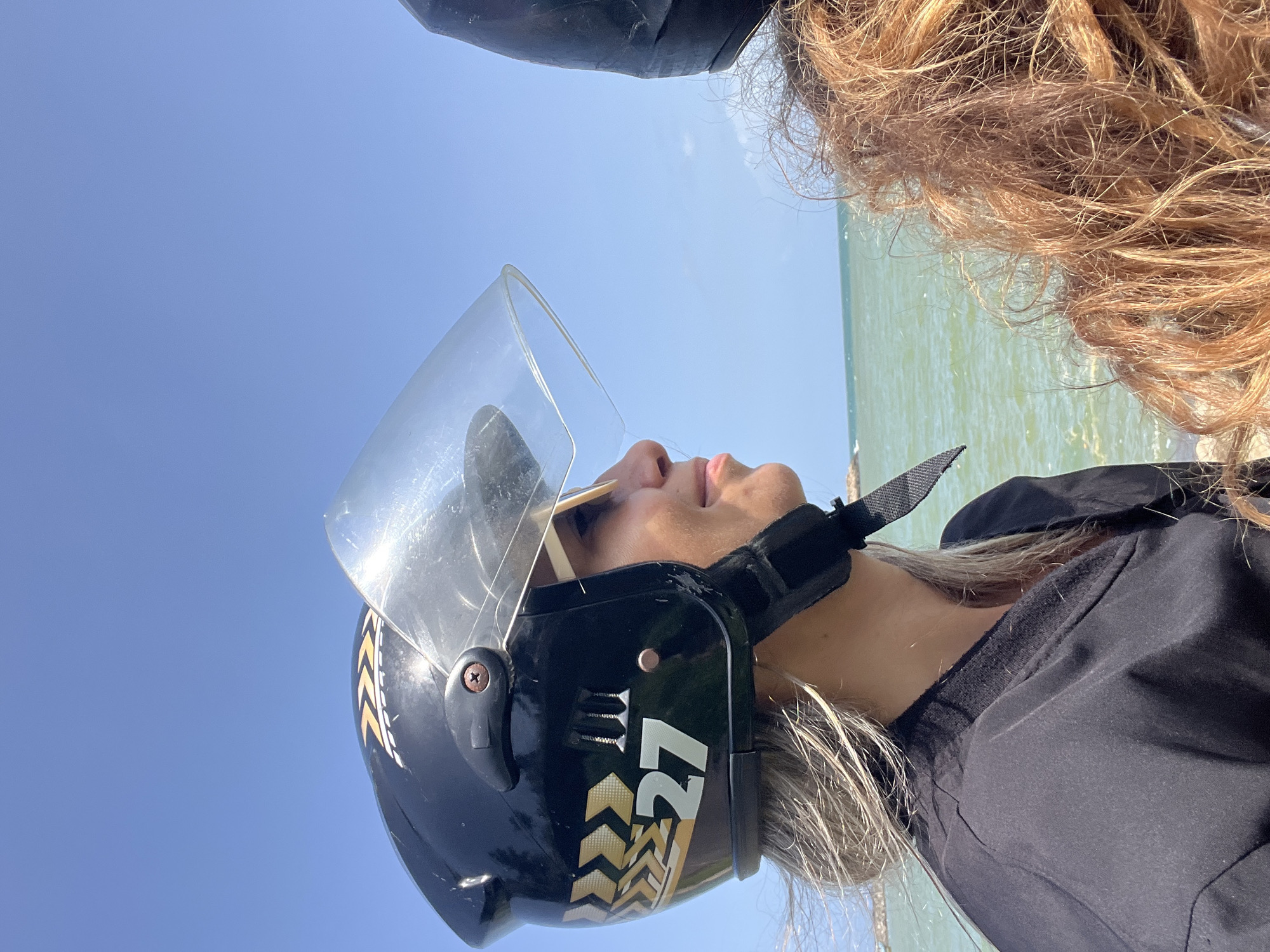 A close-up, side-profile shot captures a woman wearing a black helmet with a clear visor and gold accents, including the number "27." She has blonde hair, is wearing sunglasses, and is dressed in a black garment. Her skin appears tanned. Next to her, on the right side of the frame, is another person, partially visible, with reddish-brown curly hair. They are both looking towards the right of the image, with a body of water and a clear blue sky in the background. The water is a greenish-blue color, and the sky is cloudless. This scene is set in Dondra, Sri Lanka, likely near the coast given the presence of the sea.