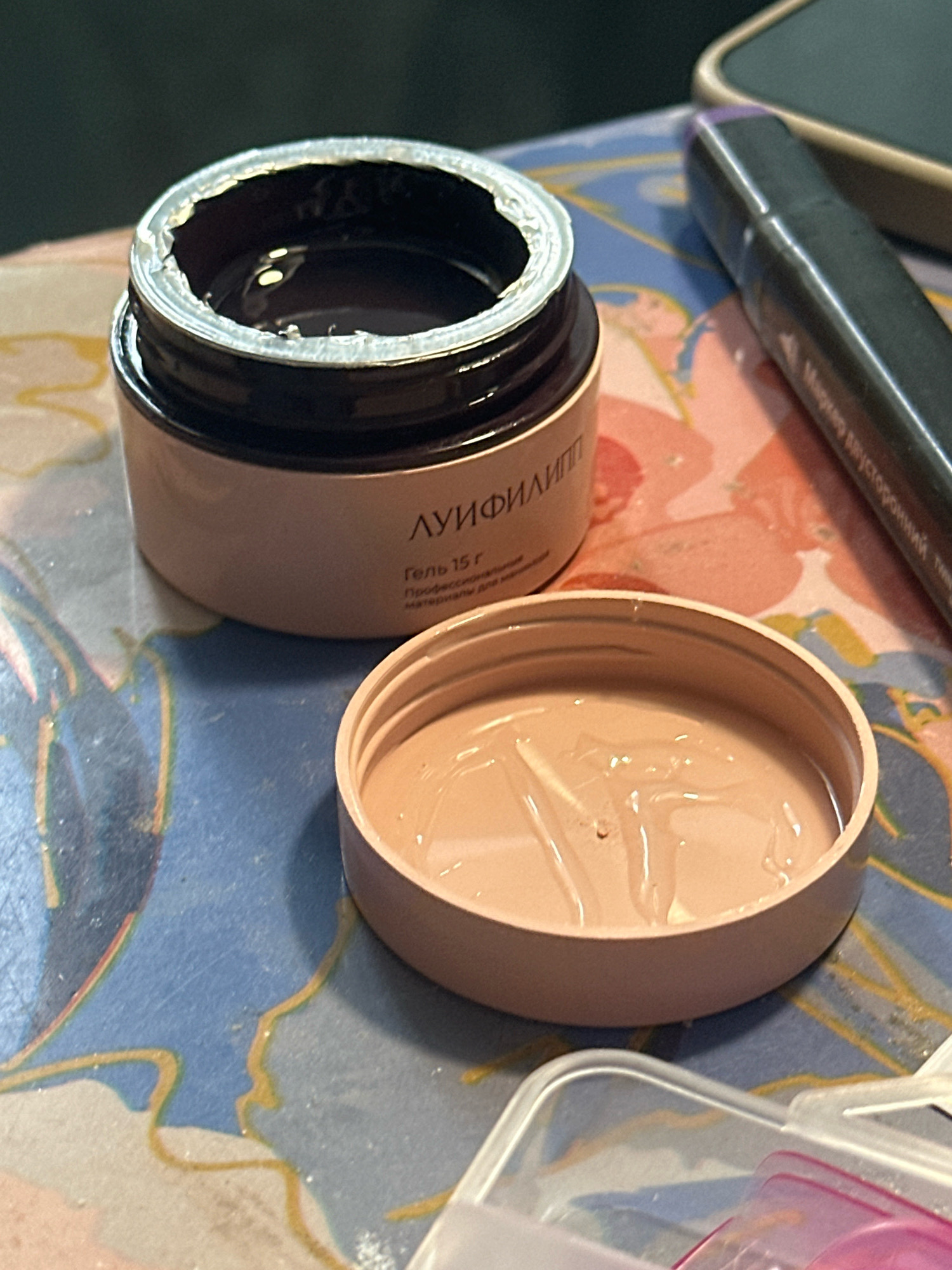 An overhead, eye-level shot shows an opened jar of dark brown substance with a tan lid off to the side, revealing a beige gel inside. The jar sits on a surface covered with a blue, pink, and yellow abstract floral patterned cloth. A black marker with a purple tip is positioned to the right of the jar. A portion of a light-colored object, possibly a phone or tablet, is visible behind the marker. A clear plastic container with pink accents is in the foreground at the bottom of the frame. The scene is indoors and well-lit.