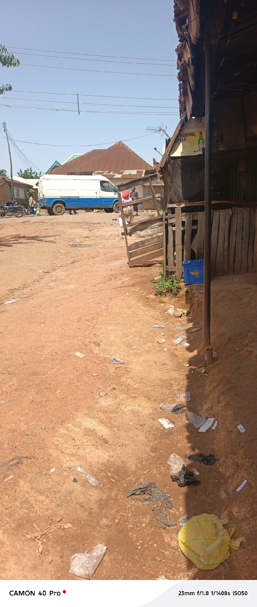 The image depicts an outdoor scene in Barikin Gidar Kanat, Nigeria. The foreground is dominated by a dirt road covered in debris, including plastic bags and other refuse. To the right, a rustic wooden stall with a corrugated metal roof is visible, with a blue plastic crate on the ground in front of it. Further into the scene, a white van with a blue stripe is parked on the dirt road. Several people are present, with two individuals standing near the van and another person, possibly a woman with a red head covering, standing to the right of the van. In the background, various buildings with corrugated metal roofs are scattered across the landscape, under a clear blue sky. Power lines are strung across the upper portion of the image. The overall impression is one of a dusty, unpaved area with signs of commercial activity and general litter.