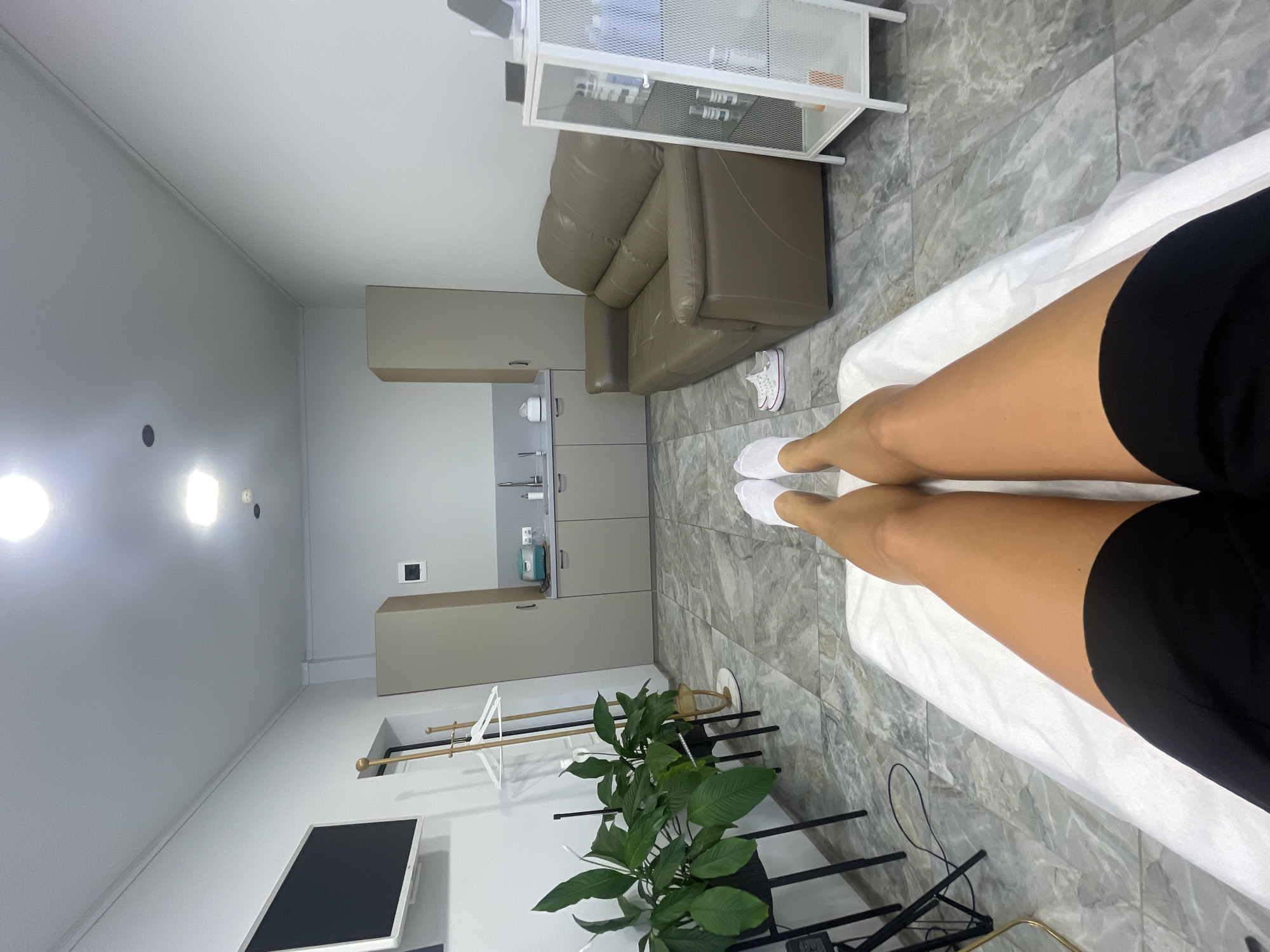 This image presents an indoor scene, likely a modern treatment room within a medical office, beauty salon, or spa in Sochi, Russia. The perspective is from a person lying on their back on a white examination or massage bed, looking towards the foot of the bed and the opposite side of the room.
In the immediate foreground, the person's tanned legs are visible, clad in black shorts or a similar garment, with white disposable socks covering their feet. The bed itself is covered with a fresh, white disposable paper sheet.
The room features a neutral and contemporary design. The floor is covered with grey and white marbled tiles. Against the far wall, a built-in cabinetry unit with light grey doors and a light beige countertop incorporates a sink with a modern faucet, a wall-mounted dispenser (possibly for soap or sanitizer), and various small bottles and containers on the shelves, suggesting a functional area.
To the right of the cabinetry, a comfortable-looking grey leather armchair or small sofa is positioned on the tiled floor. Next to it, a pair of white sneakers with pink accents are neatly placed. Above the armchair, a white metal shelving unit holds numerous small products, reinforcing the impression of a professional treatment space.
Further left in the room, a large green potted plant, resembling a Peace Lily, sits on the floor, adding a touch of natural color. A black stand with horizontal bars is also visible near the plant, alongside a partial view of a wall-mounted flat-screen television in the bottom left. The walls are light grey, and the ceiling is white, featuring several bright light fixtures. A light brown, rectangular box-like structure, possibly an air conditioning unit or vent, is mounted high on one wall.
The room is brightly illuminated by artificial lighting, creating a clean, organized, and functional ambiance. The person's posture suggests they are relaxed, likely preparing for or undergoing a procedure or treatment. No visible text is discernible in the image.