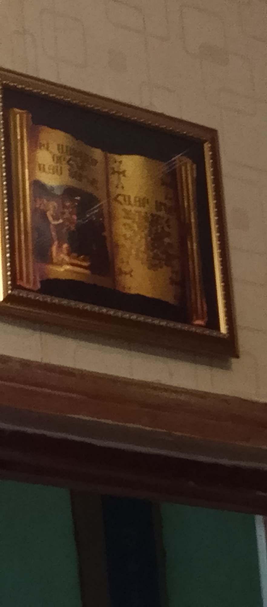 An ornate framed painting hangs on a textured wall. The painting depicts an open scroll with text and illustrations. On the left side of the scroll, a scene shows several figures, possibly religious in nature, with one figure appearing to be kneeling. The scroll has decorative columns on either side, framing the text and imagery, which appears to be in a script that is difficult to decipher. The colors in the painting are predominantly gold, brown, and darker tones for the figures and background. The frame is a dark wood with a decorative pattern along its inner edge. Below the painting is a wooden shelf or ledge. The overall scene is an indoor shot, with the wall texture and lighting suggesting it is a domestic or institutional setting. The city of Unknown, Unknown is mentioned in the prompt, but there is no visual information within the image to corroborate this location.