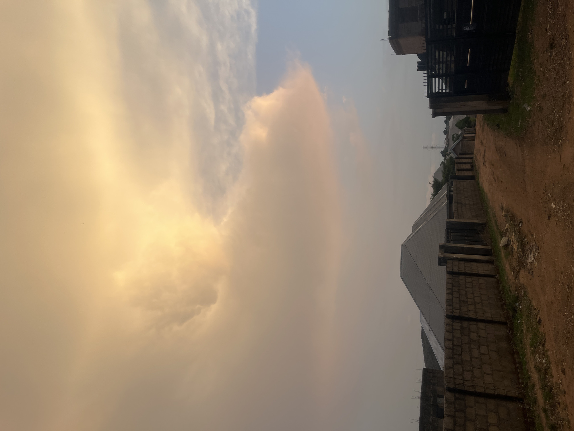 The image depicts a dirt road in Barakin Akawo, Nigeria. The road is bordered by buildings and walls. The sky is filled with clouds, with a golden hue. In the distance, there is an electrical tower. A metal gate is visible on the top right corner, attached to a building on the right. The buildings are a mix of finished and under construction.