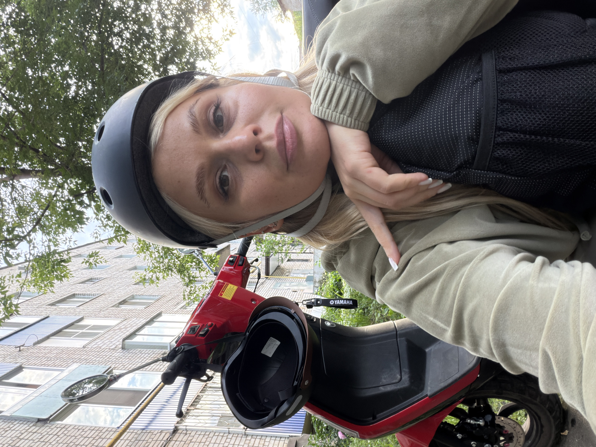 In Sarapul, Russia, a young woman with blonde hair and a helmet is captured in a selfie. She is positioned next to a red electric scooter, with a brick building and lush greenery forming the backdrop. The scooter, a Yamaha model, has its helmet secured to the front, and a distinctive Yamaha keychain hangs from the handlebars. The woman's attire consists of a light green hoodie, and her hand rests on a black backpack. The lighting suggests it is daytime, with sunlight filtering through the trees, casting a warm glow on the scene.