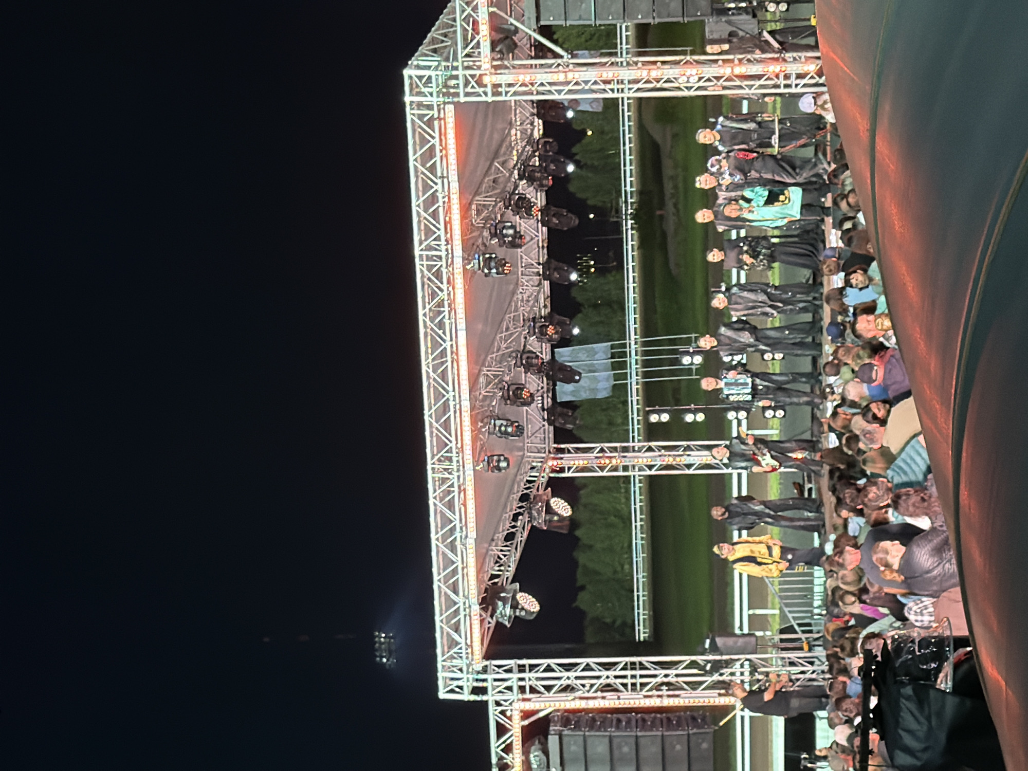 An outdoor nighttime concert stage is set up with a crowd of people gathered in front of it. The stage has a large metal truss structure with lights and speakers. Several performers, dressed in dark, stylish outfits, are on stage. One performer in the center is holding a microphone and appears to be singing or speaking. Others are playing instruments, including a guitar and an accordion. The audience is a mix of adults and some children, visible in the foreground and mid-ground. The overall environment appears to be a clear night with a dark sky above. The background shows some trees and a distant structure that resembles a tower.