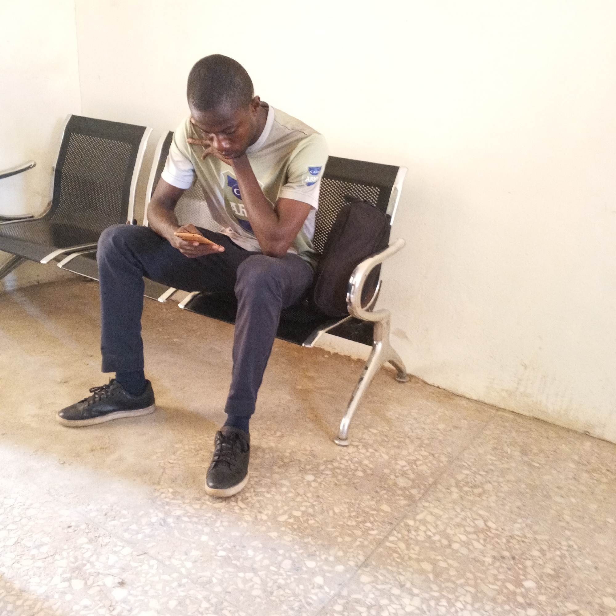 A young Black male is seated on a black mesh bench with chrome legs in what appears to be a waiting area. He is wearing a light-colored t-shirt with a logo, dark pants, and black sneakers. The male is looking down at a mobile phone in his right hand, held in his lap. His left arm rests on his left leg, with his chin on his left hand. A black bag is behind him on the bench. The floor is tiled with a terrazzo pattern, and the wall behind the bench is a light neutral color. The lighting suggests an indoor environment, possibly a public building or office. This scene is set in Arawa, Nigeria.