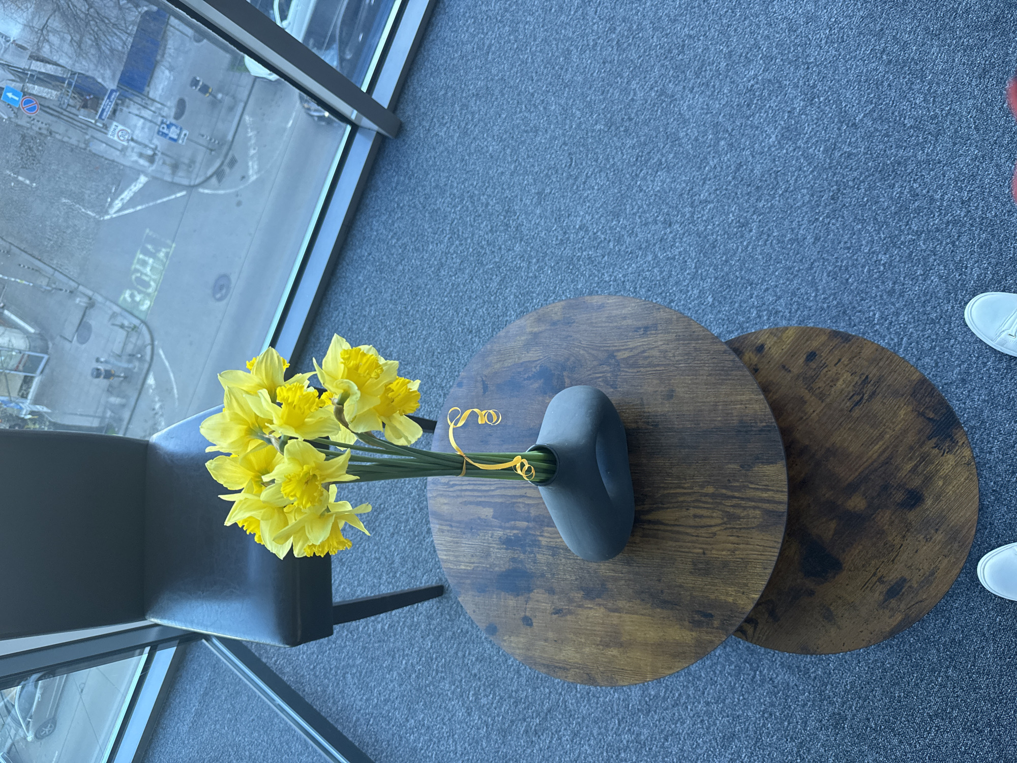 The image shows an indoor scene, likely an office space, featuring a view of a city street through a window. The street appears to be located in Sofia, Bulgaria, identifiable by Cyrillic script on street signs. A gray chair is positioned near the window, next to two round, wooden-patterned side tables. On the larger table sits a gray vase holding a bouquet of yellow daffodils tied with a yellow ribbon. Part of a person's legs and white shoes are visible on the right side of the frame, suggesting someone is standing nearby. The floor is covered with gray carpet.