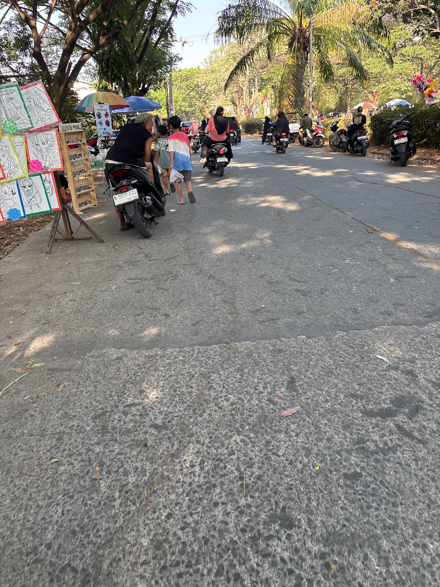 A street scene in Telagamurni, Indonesia, features a busy road with numerous motorbikes, some with passengers. On the left side of the road, a stall displays drawings and coloring pages for sale, with a wooden stand holding several small items, possibly toys or trinkets. A person is seated on a motorbike in front of the stall, and two children are standing nearby. The road is paved and appears to be in moderate condition. Trees line the sides of the road, casting dappled shadows on the asphalt. In the background, more motorbikes are visible, as well as people and several umbrellas, suggesting a market or gathering. Palm trees are also present, indicating a tropical or subtropical climate. The overall impression is one of a lively, everyday scene in a populated area.