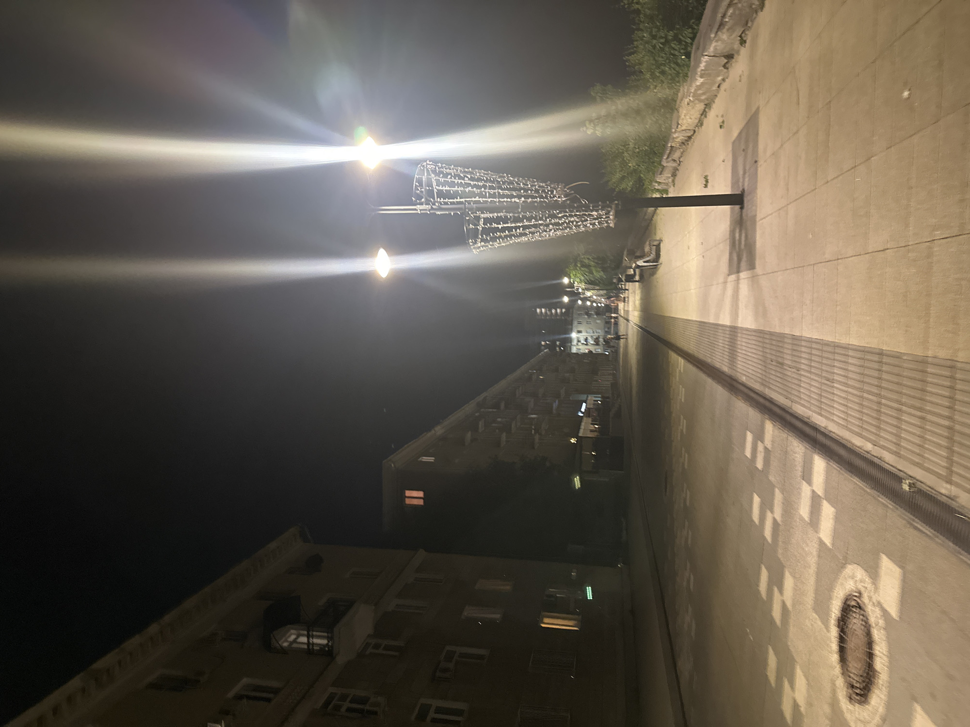 The image depicts an empty pedestrian walkway at night in Saratov, Russia. The walkway is illuminated by streetlights, casting long beams of light across the scene. On the left, a multi-story apartment building with balconies is visible. The walkway itself is paved with light-colored tiles and features a patterned design in some sections. A manhole cover is present on the walkway near the bottom left. To the right of the walkway, there is a low wall bordering an area with trees and bushes. A lamppost with a decorative, string-light adorned structure stands prominently on the walkway. The sky is dark, and there is a slight haze or fog present, contributing to the glow of the lights. The scene appears serene and undisturbed, with no individuals or active events occurring.