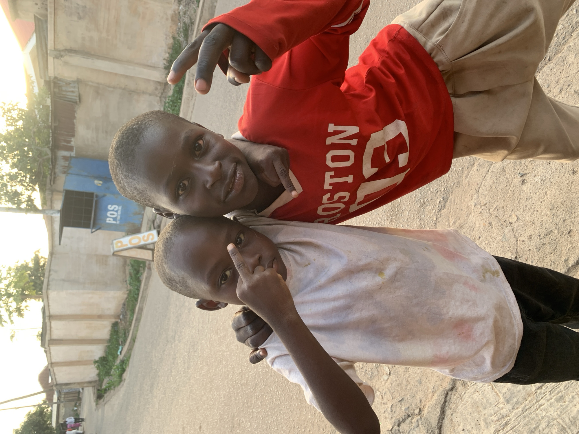Two young boys stand closely together on a paved street in Jos, Nigeria, posing for the camera. The boy on top wears a red shirt with "BOSTON" printed on it and beige pants, making a peace sign with his fingers. The boy below wears a dirty white t-shirt and dark pants, extending his middle finger toward the camera. A building with a "POS" sign is visible in the background, indicating a point-of-sale location, common for financial transactions in Nigeria. The street appears to be residential with vegetation and additional buildings visible. The lighting suggests either early morning or late afternoon.