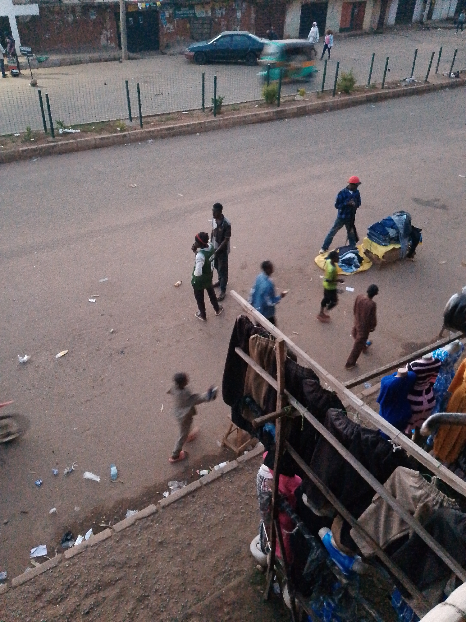 The image shows a street scene in Bukuru, Nigeria, with several people visible. In the foreground, there is a wooden structure with various items of clothing and fabric draped over it, suggesting a market or stall. The street is paved with asphalt and there are some scattered pieces of debris.
Several individuals are walking or standing on the street. In the middle ground, one person wearing a red cap and a blue checkered shirt is walking. Further back, there are two people standing together, one wearing a green vest. A child is visible running in the middle of the road. In the background, there is a dark-colored car on the road, and a blurred green and yellow vehicle that appears to be a tuk-tuk or tricycle. Buildings line the street in the background, with some visible posters or signs on their facades. A metal fence runs along the side of the road, separating it from a dirt path with some sparse vegetation.
