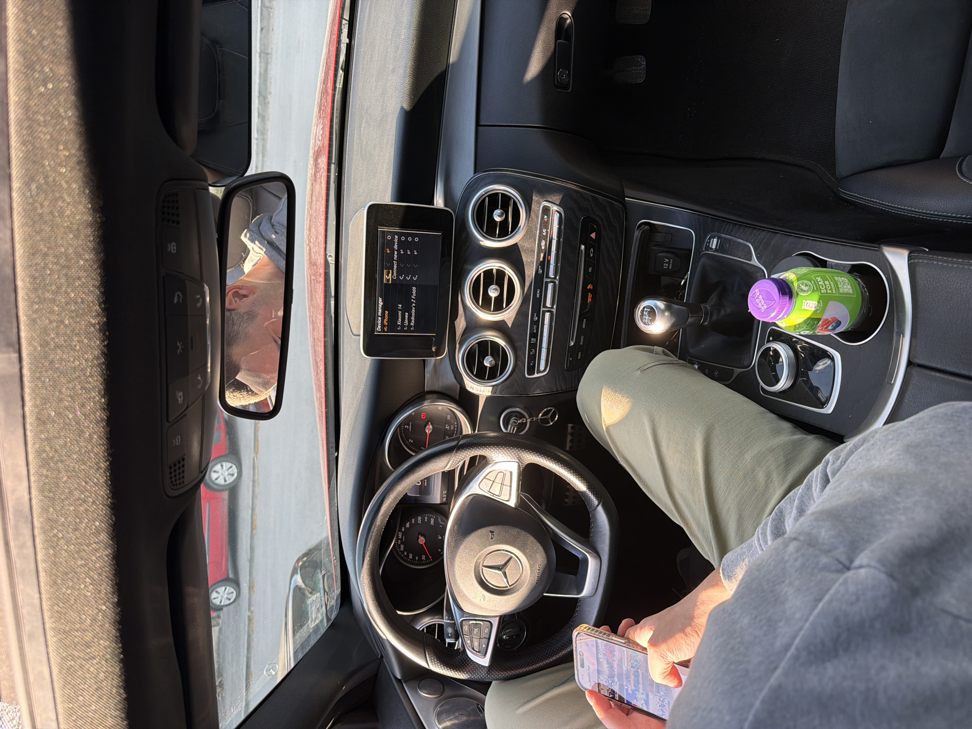 The image shows the interior of a Mercedes-Benz car. The driver is visible in the rearview mirror, and another car is visible outside the window. The car's dashboard is visible, with a display screen showing options for connecting to devices like iPhone, Xiaomi 14, Цанев, and Radoslav's Z Fold6. The steering wheel has the Mercedes-Benz logo. The driver is holding a smartphone in his hand, and there is a bottle of juice in the cup holder. It is unknown if this image was taken in Varna, Bulgaria. There is no information in the image to substantiate this.