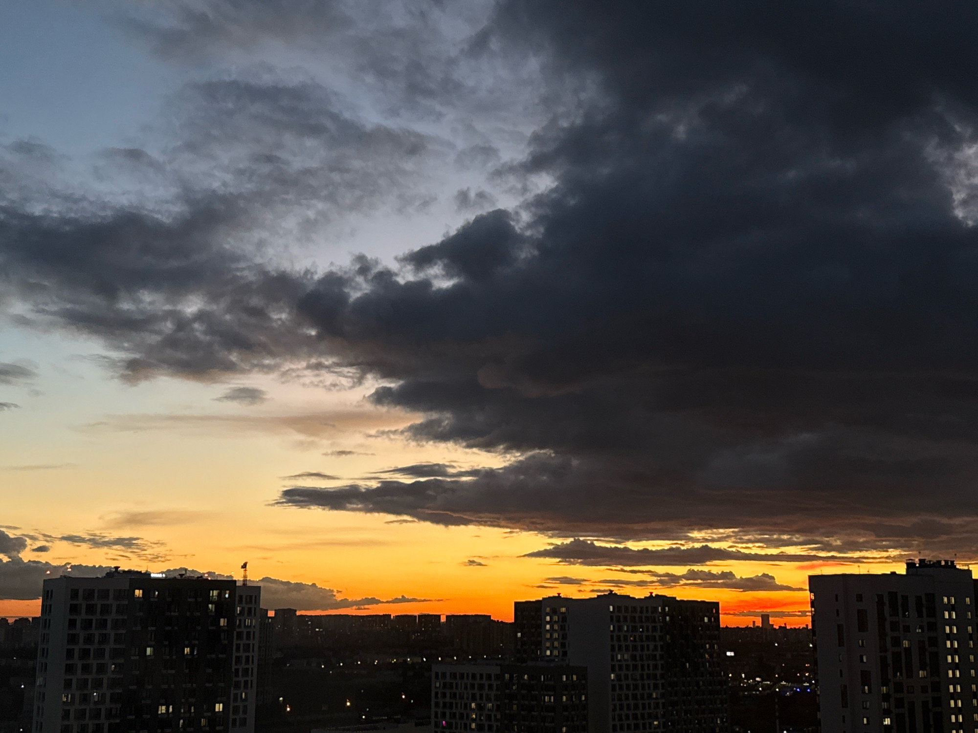 The image depicts a dramatic sunset over the city of Tomilino, Russia. Dark, ominous storm clouds dominate the upper portion of the sky, transitioning into lighter grays and blues towards the horizon. Below the clouds, a vibrant band of orange and yellow light stretches across the sky, indicating the setting sun. Silhouetted against this colorful sky are the dark outlines of numerous high-rise apartment buildings, typical of a modern urban landscape. The windows of some buildings are illuminated, suggesting that the photo was taken in the late evening. The foreground is occupied by the tops of these buildings, with one in the lower left corner showing distinct architectural details and multiple floors with windows. In the distance, the city extends into the horizon, with more buildings and possibly industrial structures visible. The overall scene conveys a sense of atmospheric drama and the encroaching darkness of nightfall.