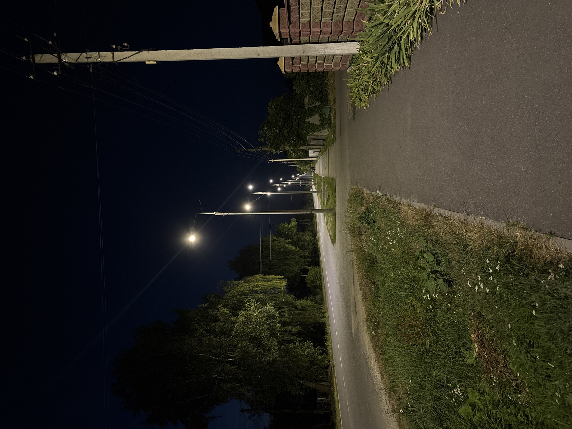 The image depicts a straight, paved road at night in the city of Lanivtsi, Ukraine. The road is lined on the right by a sidewalk and a brick fence. On the left side of the road, there is a grassy verge with trees. Numerous streetlights illuminate the scene, casting a yellow glow on the road and sidewalk. Power lines are visible stretching across the dark night sky, supported by utility poles. The atmosphere is calm and quiet, with no people or vehicles present.