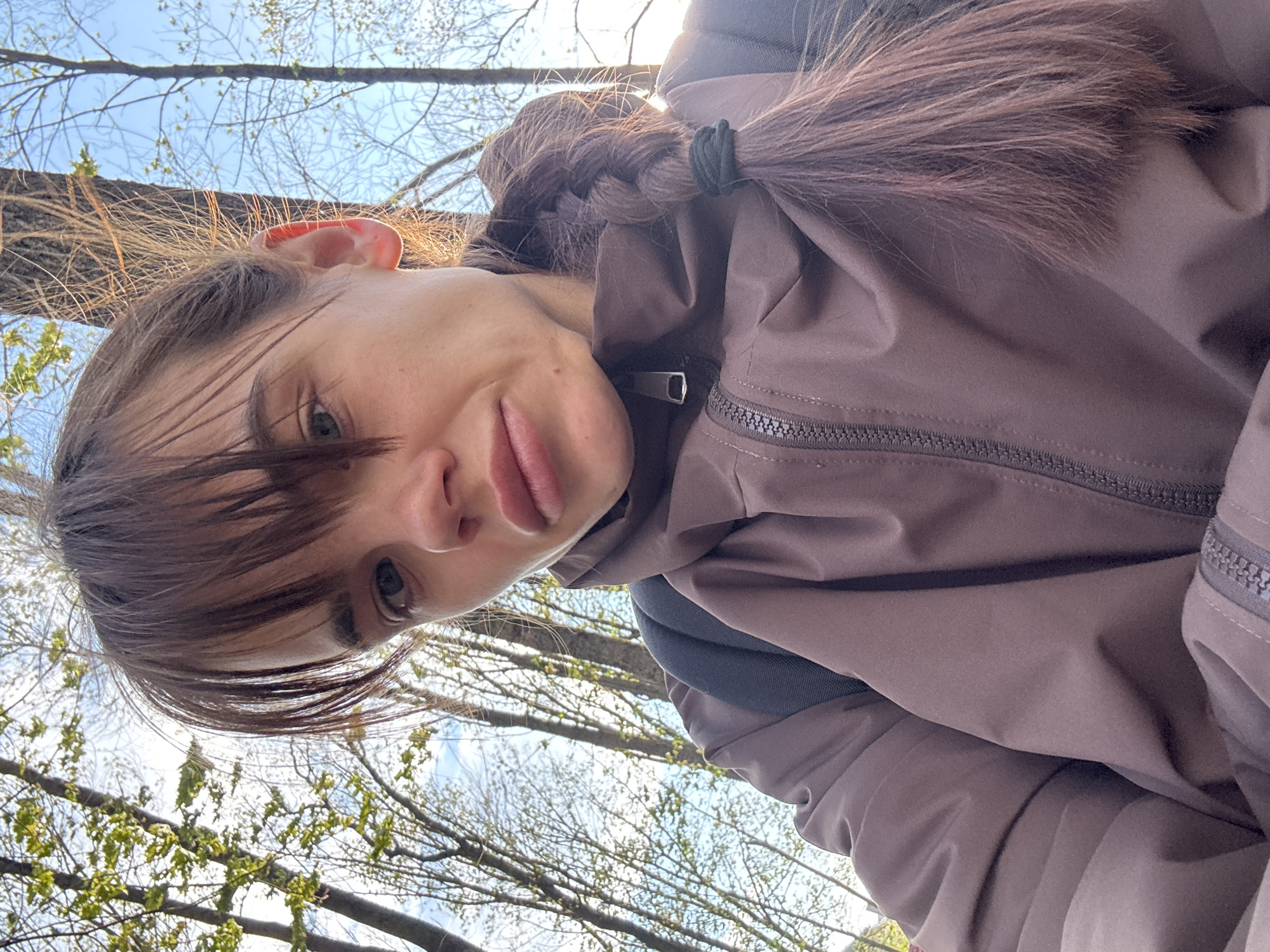 This medium close-up portrait depicts a young woman taking a selfie outdoors in Rostov-on-Don, Russia. The image is oriented sideways, with the woman's head tilted significantly to the left, as if she is looking up at the sky from a low angle. Her face, including her right eye, nose, and prominent lips, is visible, framed by light brown bangs across her forehead. Her brown hair is styled in a long braid, secured with a black elastic hair tie, and catches the sunlight. Her right ear is partially visible, showing translucence from the light source behind her.
She is wearing a brown or taupe-colored jacket with a zippered front and what appears to be a collar or hood. A dark gray or black strap, possibly from a backpack, is visible over her right shoulder. The background features a bright, clear blue sky with some wispy white clouds. Numerous tree branches, mostly bare but with nascent small green leaves and buds, crisscross the upper portion of the frame, indicating an early spring setting. The sunlight is strong, creating highlights on her hair and illuminating the branches. Her expression is neutral, with her gaze directed towards the camera. There is no visible text in the image.