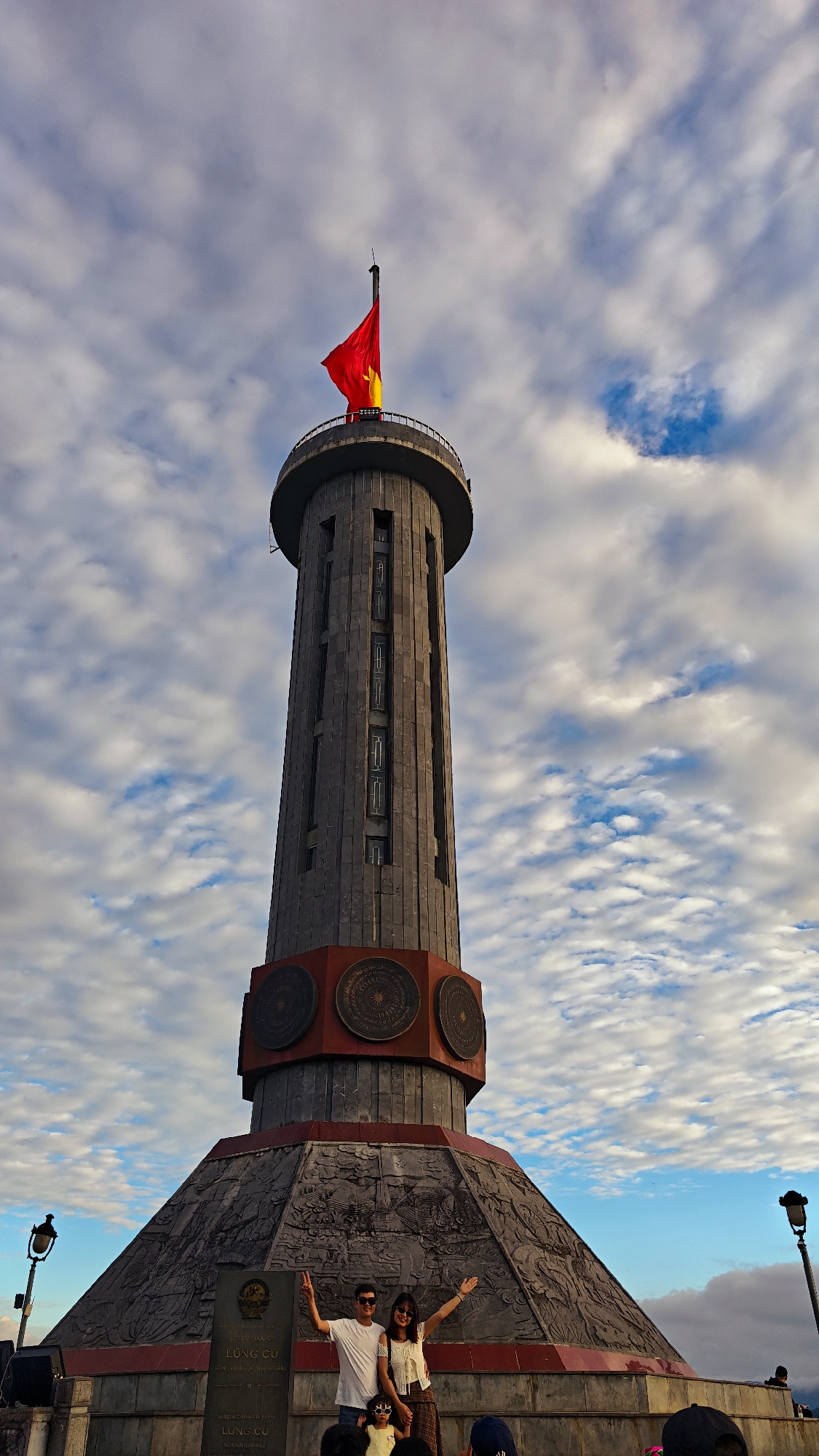 Here's a detailed description of the image:
The photograph captures the Lung Cu Flag Tower in Lũng Cú Commune, Vietnam. The tower is the main focal point, towering against a backdrop of a sky filled with puffy, white clouds scattered across patches of blue. At the tower's peak flies the flag of Vietnam, brightly colored in red and yellow.
The structure of the tower consists of a cylindrical shaft, divided into sections with decorative bronze-colored elements. The base expands in a conical form, etched with detailed carvings and inscriptions.
In the foreground, at the base of the tower, a group of people are standing. A man and a woman, seemingly a couple, are posing with their arms raised in a gesture of enjoyment or celebration. Others are positioned in front of them with sunglasses and what looks like an eager attitude.
The overall scene has a touristic feel, documenting the experience of visitors at this landmark location.