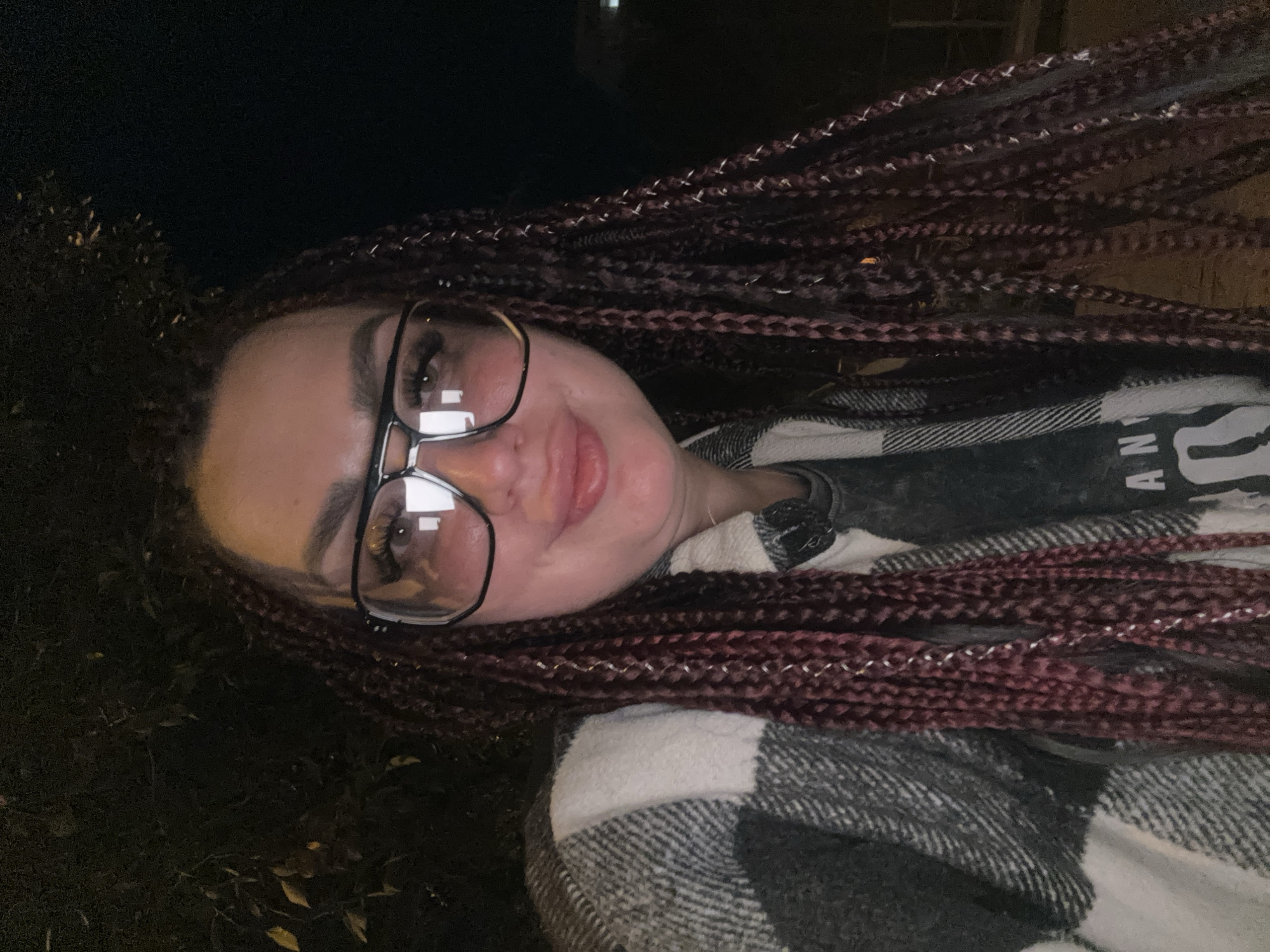 A medium close-up photograph shows a woman with dark braided hair and large, dark-rimmed glasses. She is wearing a checkered shirt over a dark t-shirt with some text on it. The background is dark and appears to be outdoors at night, with some foliage visible. The lighting is dim. The woman is looking directly at the camera. This image was taken in Tolyatti, Russia.