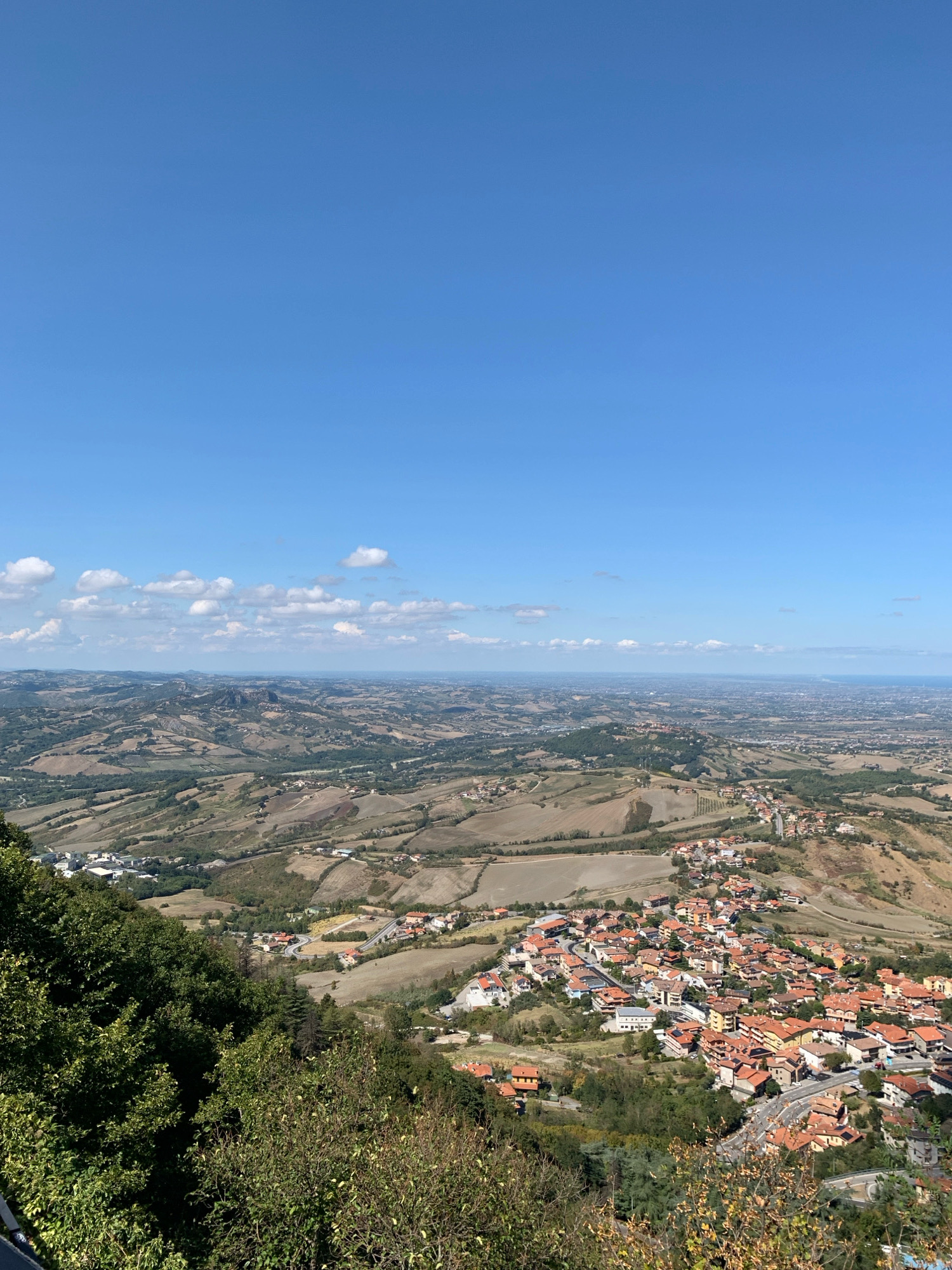 This is a high-angle, wide shot of the city of San Marino, San Marino, taken from a vantage point on a hill. The foreground is dominated by lush green foliage, with some dry, brown vegetation interspersed. Below, the city unfolds across the rolling hills, characterized by its terracotta-roofed buildings clustered together. Winding roads are visible, connecting different parts of the settlement. The landscape extends into the distance, revealing a vast expanse of countryside with fields and more scattered settlements, eventually meeting the hazy horizon. The sky above is a clear, bright blue, with a few scattered white clouds. The scene depicts a sunny day, with excellent visibility over the cityscape and surrounding terrain. No people are visible in the image.