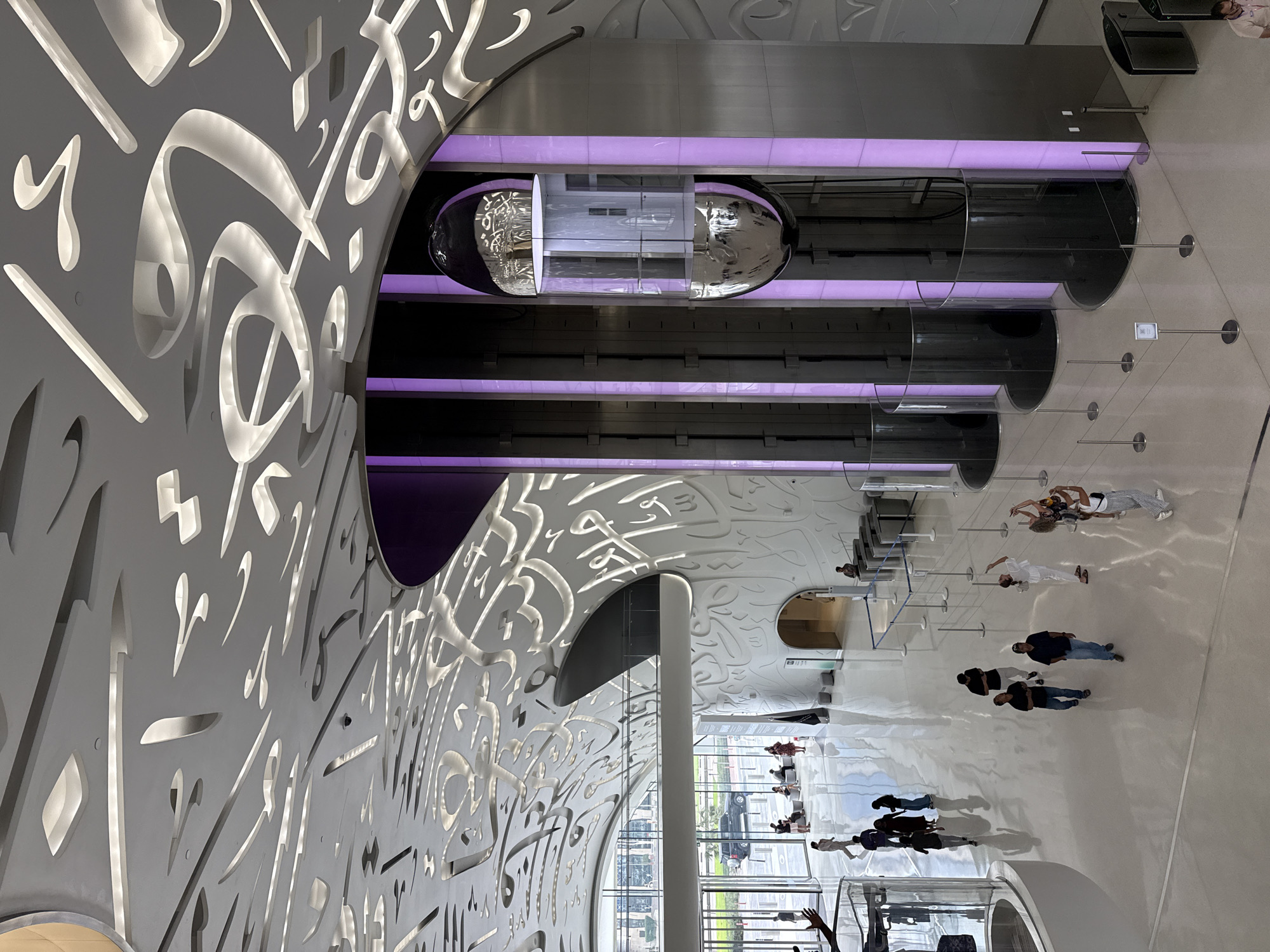 The image depicts the interior of the Museum of the Future in Dubai, United Arab Emirates. The museum's distinctive elliptical structure, adorned with Arabic calligraphy, is visible. The interior features a multi-level design, with glass-enclosed elevators or pods highlighted by purple lighting. Several people are present on the museum floor, seemingly observing the architecture and exhibits. The environment is modern and futuristic, with a focus on innovative design elements and a bright, open space.