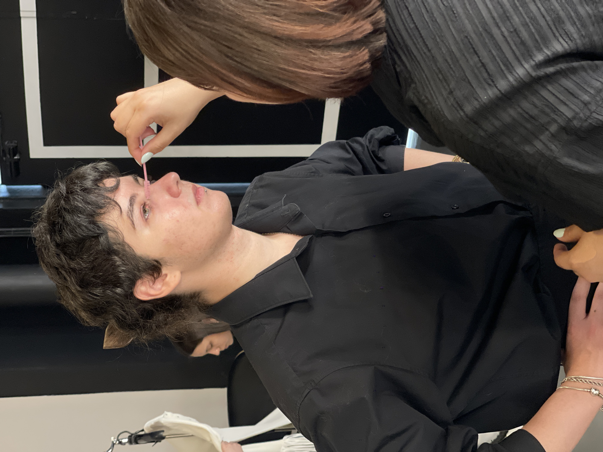 In Kyiv, Ukraine, a close-up shot captures a person receiving makeup application. The subject, with curly dark hair and a piercing on their lip, is wearing a black button-up shirt. Another person, with short brown hair, is applying pink mascara to the subject's eyelashes. The background features a black wall with a white rectangular outline, suggesting an indoor setting, possibly a studio or backstage area at an event. The lighting is bright, illuminating the faces of both individuals. The scene conveys a sense of preparation or backstage activity.