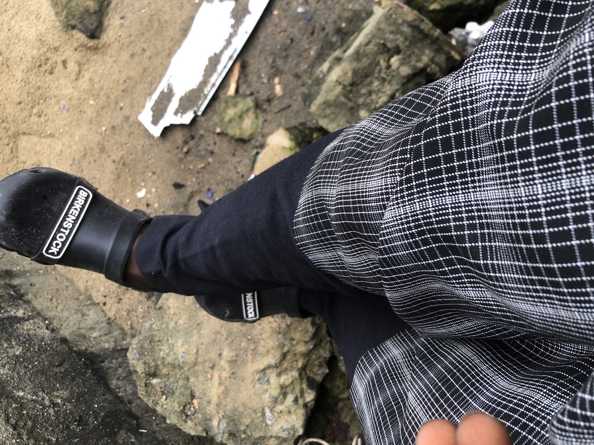 The image is a close-up shot of a person's legs and feet, wearing black Crocs with the "BIRKENSTOCK" logo visible on the strap, and dark pants. The pants have a black and white plaid pattern. The person is sitting on a rough, textured surface, likely rocks and sand. In the background, there is debris on the sandy ground, including a white, rectangular object. The overall environment appears to be outdoors, possibly near a body of water or a construction site. The lighting suggests it is daytime. The location is identified as Ajah, Nigeria.
