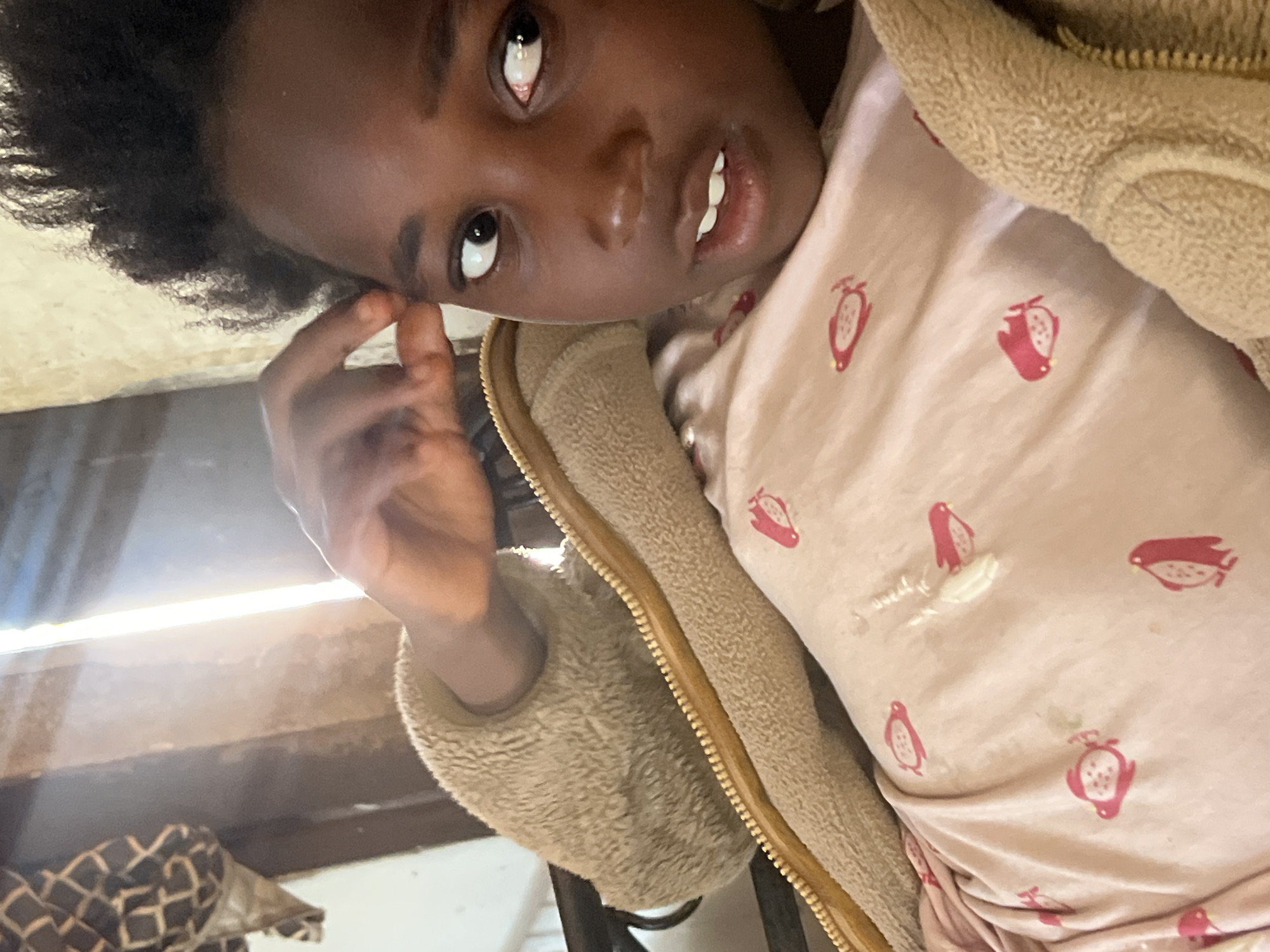 A young Black girl with dark, curly hair is lying down, looking at the camera. She is wearing a light pink shirt with a pattern of red penguins and a fuzzy tan jacket with a yellow zipper. Her left hand is raised to her forehead. The background is out of focus but appears to be an indoor setting with natural light streaming in from the side, suggesting a room with windows or an opening. The lighting creates a bright, somewhat blown-out effect on the right side of the image. The setting does not provide specific indicators of its location within Jos, Nigeria, beyond being an indoor residential or common area.