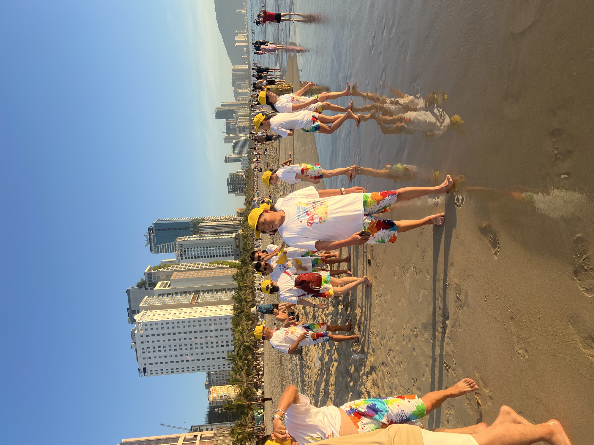 The image depicts a group of people, most of whom are wearing matching white t-shirts and colorful, tie-dye patterned shorts, along with yellow hats, on a sandy beach. The scene appears to be set in the city of Đà Nẵng, Vietnam, with a backdrop of modern high-rise buildings and palm trees characteristic of the city's coastline. The ocean water is visible in the foreground, reflecting the sky and the figures on the beach. Some individuals are walking towards the water, while others are standing and interacting. The lighting suggests it is late afternoon, with the sun casting a warm glow on the scene. There are also other people in the background, some swimming or walking along the shoreline, and distant buildings and hills are visible on the horizon.