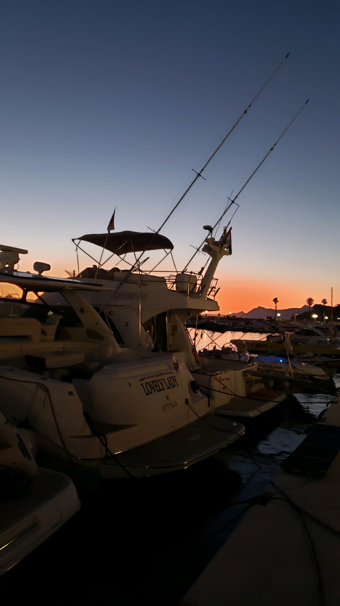 At dusk in the city of Općina Tučepi, Croatia, a white yacht named "LOVELY LADY" is docked. The yacht has two fishing rods extended from its stern. In the background, the sky exhibits a gradient of blue to orange, indicative of a sunset. Several other boats are visible in the harbor, and palm trees and hills are silhouetted against the horizon. The water reflects the fading light of the sky. No people are visible on the yacht or in its immediate vicinity.