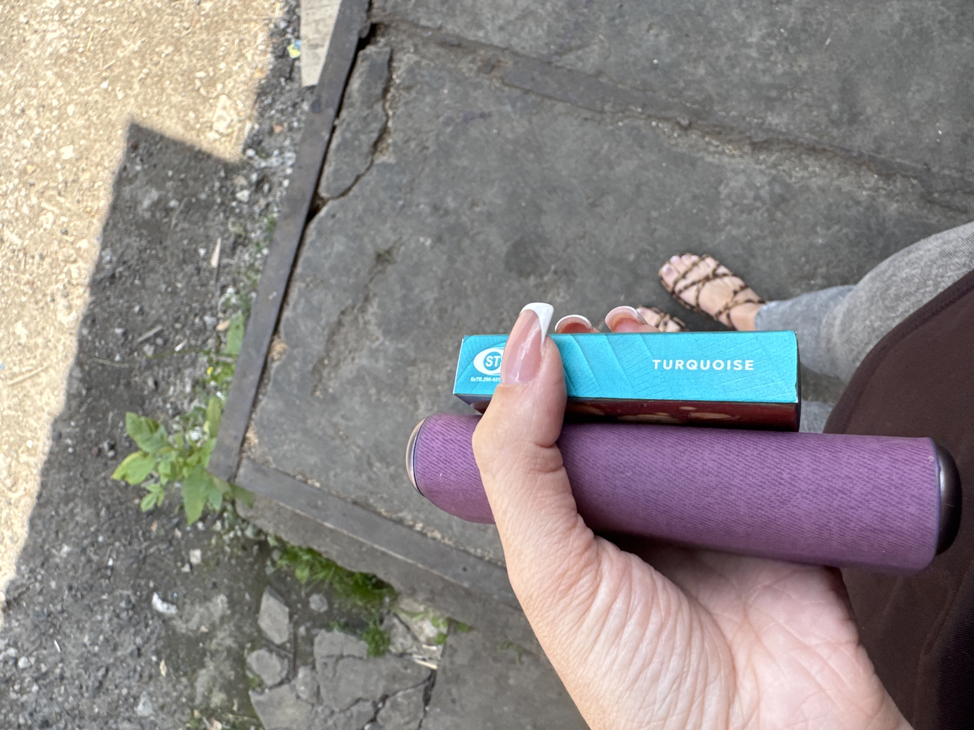 A first-person perspective shot shows a person holding a purple cylindrical object and a small, teal-colored box with white lettering that reads "TURQUOISE". The person is wearing grey leggings and brown strappy sandals. The background appears to be a cracked concrete surface with patches of dirt and sparse greenery. A shadow is cast across the ground in the upper portion of the frame.