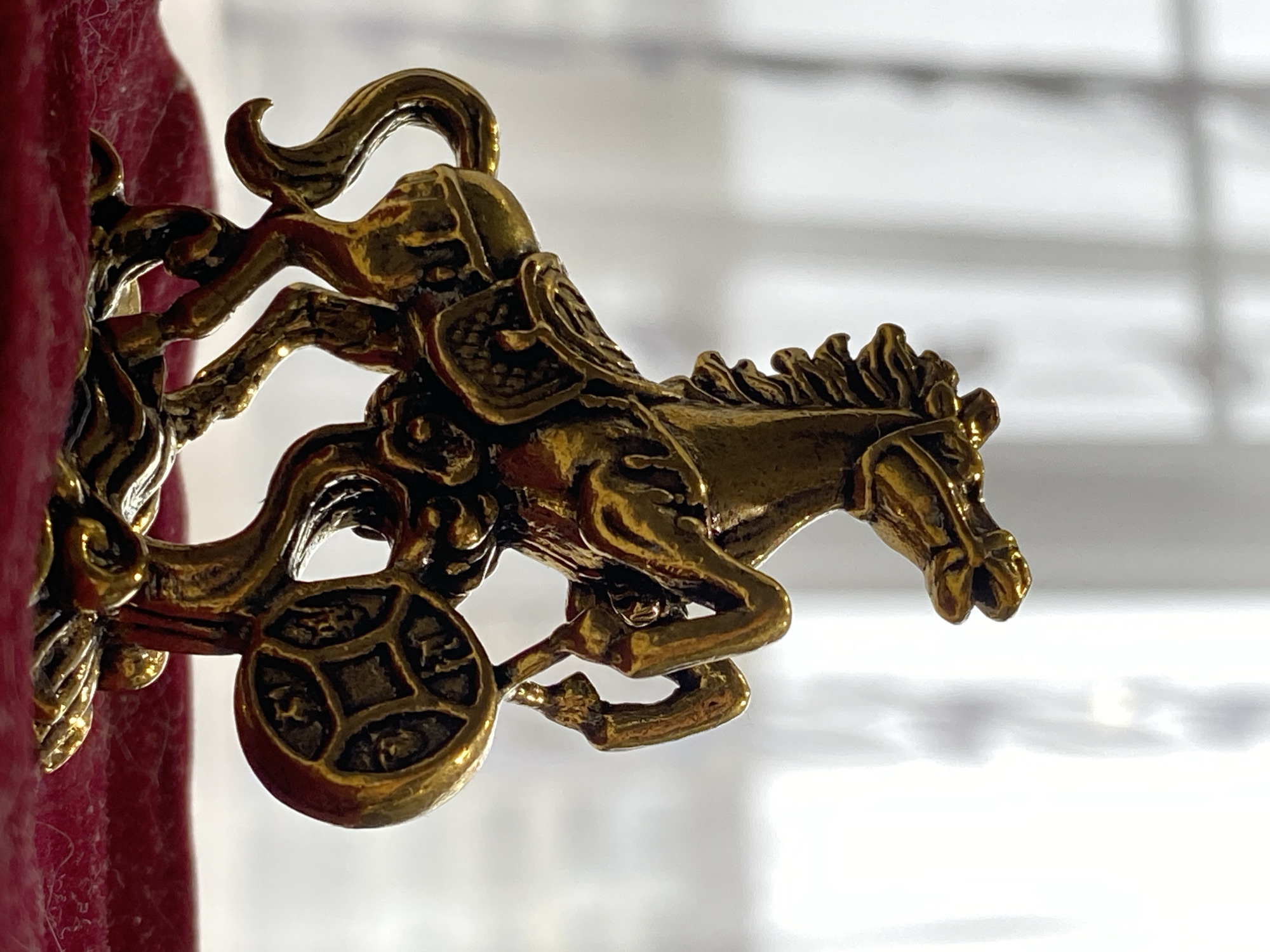 The image displays a close-up of a golden metallic figurine against a bright, blurry background and a textured, deep red surface on the left. The figurine depicts a stylized warrior or deity riding a galloping horse. The horse is captured in a dynamic pose, with a flowing mane and tail, while the rider appears to be clad in armor or elaborate robes.
A distinctive detail is a circular disc attached to the lower part of the figurine, strongly resembling an ancient Chinese "cash" coin, featuring a central square hole. Faint, stylized characters or symbols are visible around the square hole, indicative of its potential cultural origin and symbolic meaning, often associated with prosperity.
The figurine exhibits a lustrous, brass-like golden color, with intricate detailing that creates depth and catches the light. The metallic surface has a polished appearance, highlighted by reflections. The red surface provides a strong color contrast to the golden object, suggesting the figurine is placed against or attached to a decorative item or fabric. The blurred background offers no specific location cues beyond being a bright, possibly indoor setting in Markovskii, Russia. The image focuses solely on the ornamental details of this static object. No visible text is clearly legible, and no activity or emotions are present beyond the depicted movement of the horse and rider.