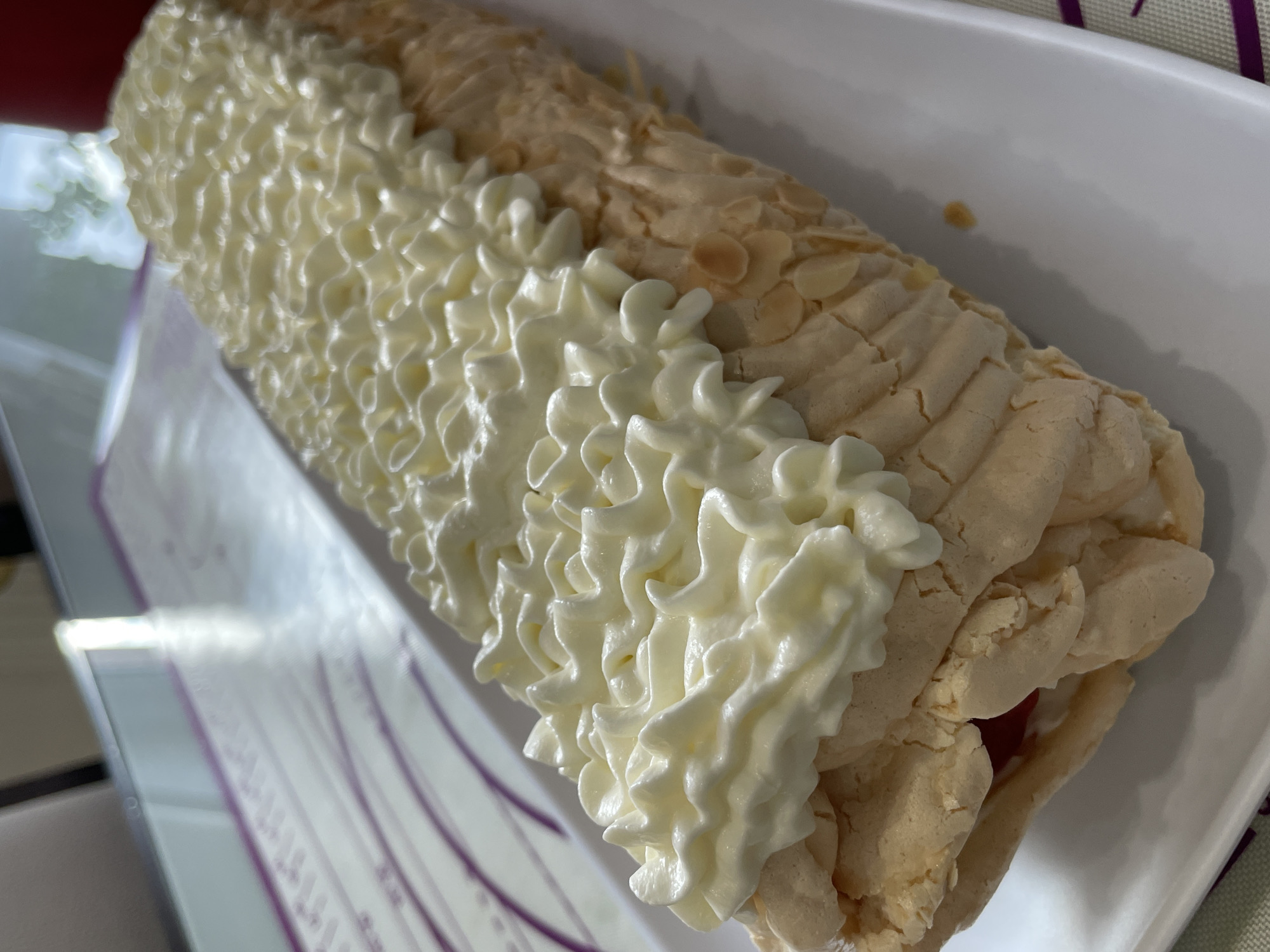 The image shows a close-up, high-angle shot of a white plate holding a pavlova roll dessert. The dessert is long and cylindrical, covered with swirled white cream on top and sliced almonds on the sides. The pavlova base appears to be light brown and textured. A glimpse of red filling is visible at one end. The dessert is positioned on a white rectangular plate. In the background, a purple and white baking mat with circular markings is partially visible, and a reflective surface, possibly a countertop or table, is also present. The scene appears to be indoors, likely in a kitchen. The photograph does not provide any specific information about the city of Saint Petersburg, Russia.