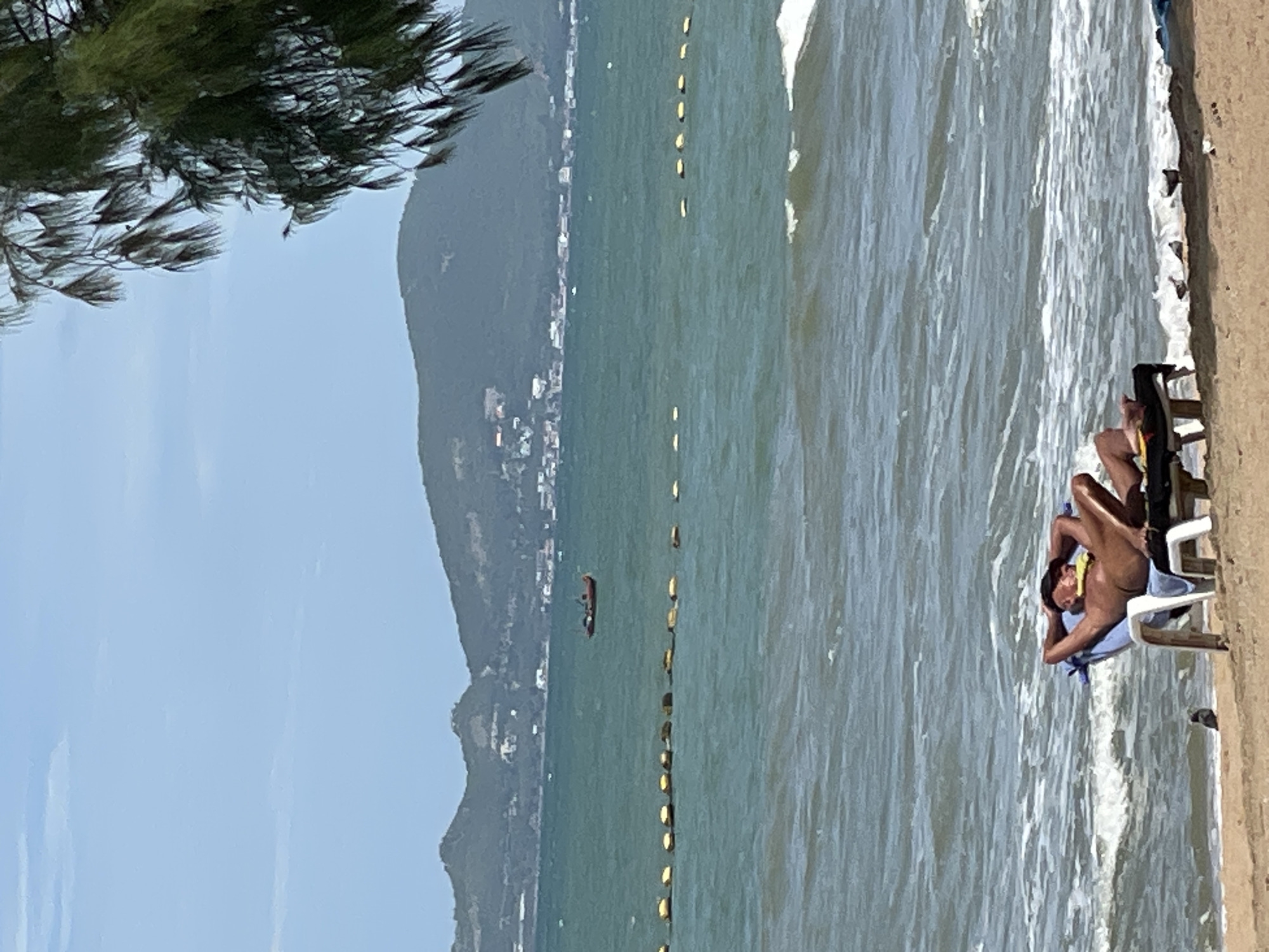 A man sunbathes on a lounge chair on a sandy beach in Pattaya City, Thailand. He is wearing a dark hat and a yellow cloth around his neck. The sea is turquoise and waves are breaking on the shore. In the distance, a line of yellow buoys stretches across the water, and a small red boat is visible near a distant shoreline dotted with buildings and green hills. A tree branch with long, green leaves hangs into the frame from the upper right corner. A pigeon stands on the sand near the lounge chair.
