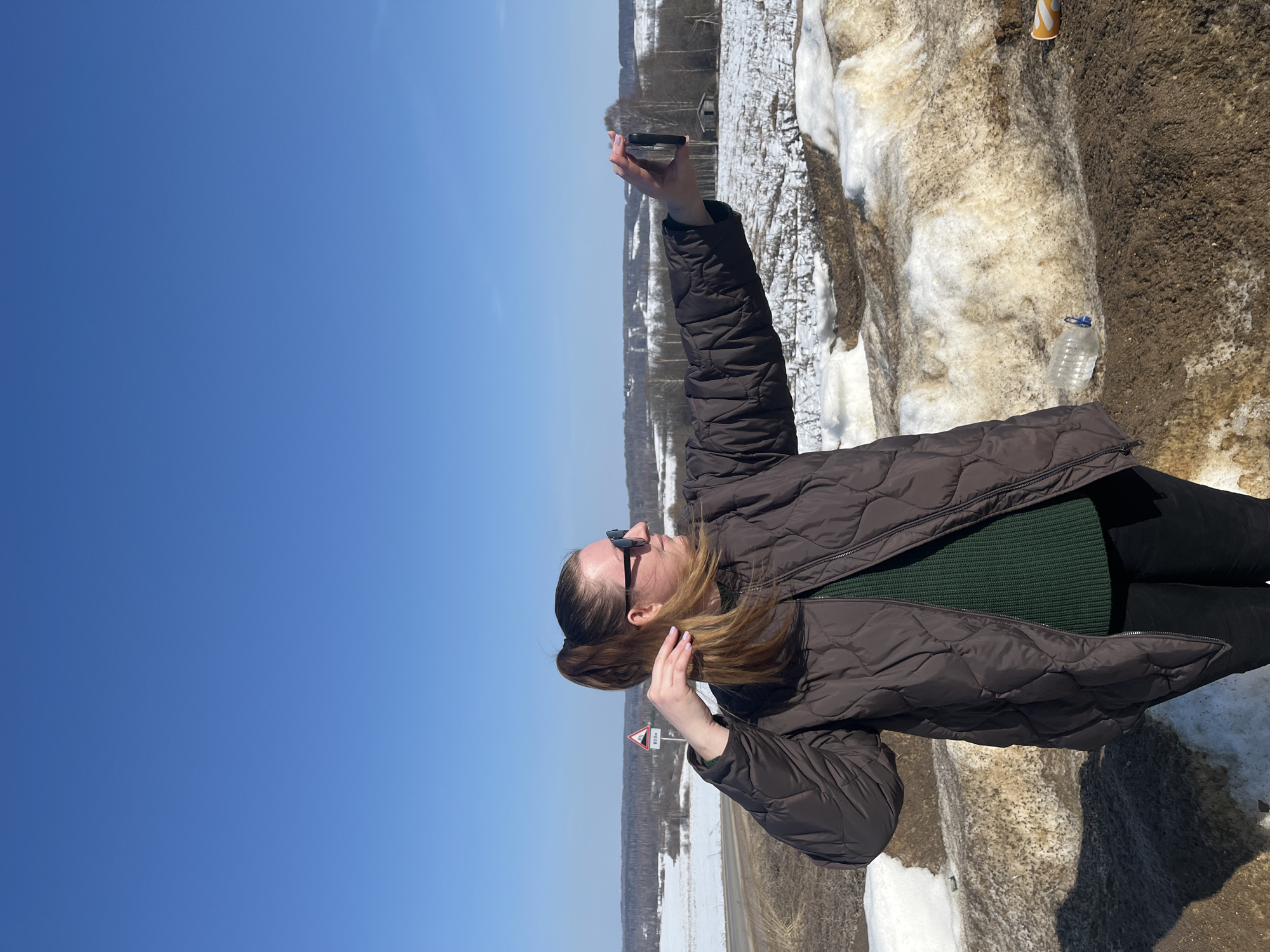 A female individual stands in a semi-rural outdoor environment near Zubovo, Russia, positioned on a mound of melting snow and brown earth next to a paved road. She has light brown hair, pulled back into a high ponytail, and wears dark sunglasses, a dark brown quilted jacket, a dark green knitted sweater, and dark pants. Her right arm is extended forward, holding a black smartphone horizontally, appearing to be taking a selfie or recording. Her left hand is raised towards her hair. Her fingernails are painted light pink.
In the immediate foreground, a clear plastic bottle with a blue cap and an orange and yellow paper cup are visible as discarded items on the ground. In the background, a red and white triangular road sign, displaying a symbol and the text "800M," is situated alongside the curving asphalt road. Beyond the road, snow-covered fields with patches of exposed brown earth extend towards a distant line of bare deciduous trees. A small, weathered wooden structure is also visible among the trees in the middle ground. The sky above is clear and bright blue, indicating sunny weather.