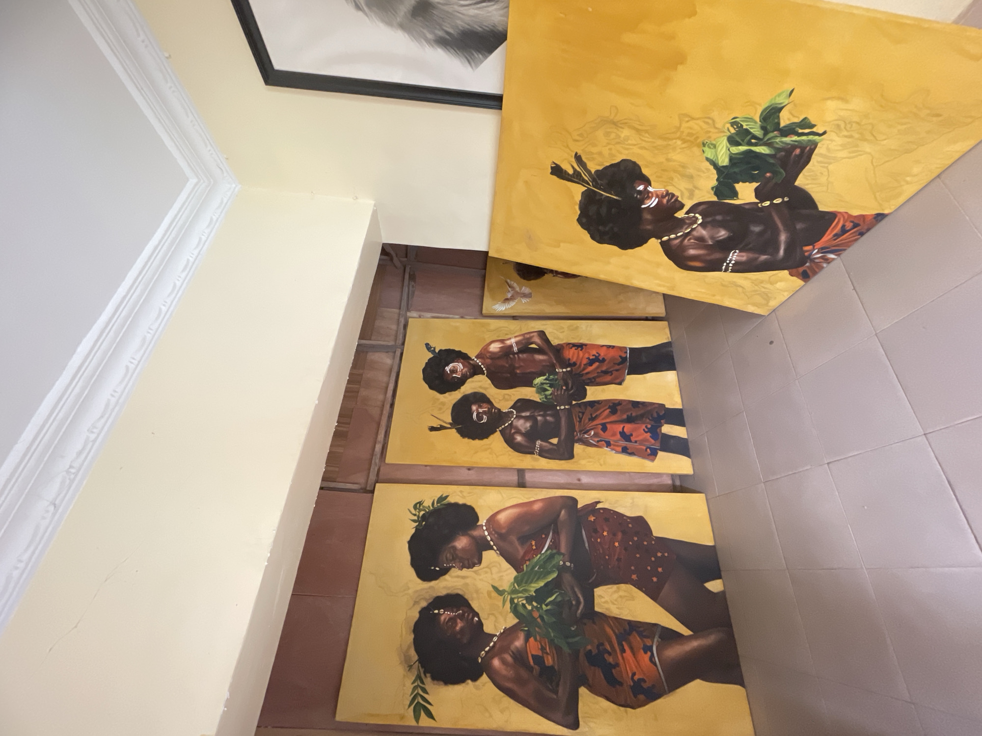 The scene captures a section of an interior space, likely a gallery or home in Ado, Nigeria, where several paintings are displayed. The paintings feature figures with dark skin, adorned in traditional clothing and jewelry, set against a yellow background. Some figures hold plants, and the paintings vary slightly in composition and arrangement of figures. The paintings are resting on the floor and leaning against the base of the wall. The floor is tiled, with neutral-colored squares. The adjacent wall is painted a light yellow. A framed black-and-white print is visible in the upper section of the frame.