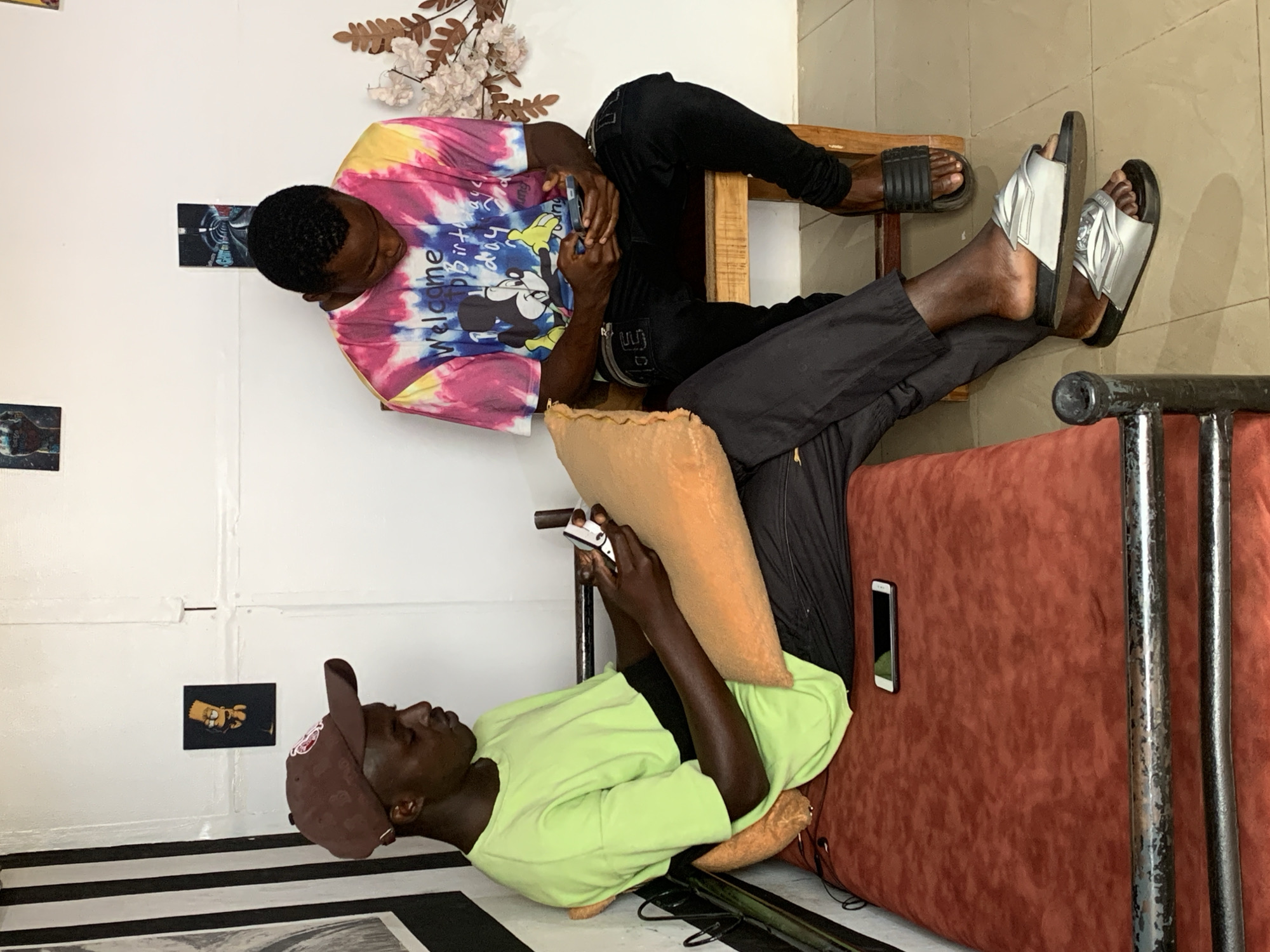 This image depicts two young Black men in an indoor setting, likely in Jos, Nigeria. The image is oriented vertically.
The man in the foreground, wearing a lime green t-shirt and a brown baseball cap, is lying down and looking at his phone. He is positioned on a rusty red sofa or bench. Next to him is a brown pillow and some cables.
The second man is seated on a wooden bench, wearing a colorful tie-dye t-shirt with a Mickey Mouse graphic and the text "Welcome to the best birthday party," and black pants. He is looking down at his phone, which he holds in his hands. He is wearing black slides on his left foot and silver slides on his right.
The background features a white wall with some framed pictures, including one that appears to be of Bart Simpson and another abstract image. There are also decorative dry flowers and branches on the wall. The lighting suggests it is daytime. The overall mood seems relaxed, with both individuals engaged with their devices.