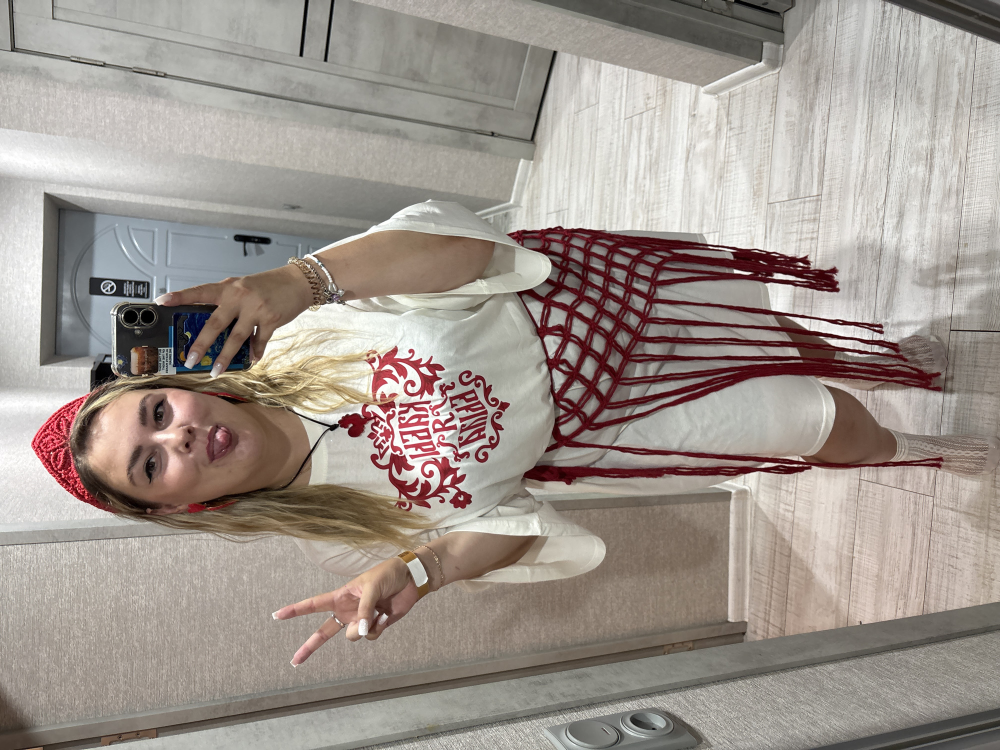 A young woman is taking a selfie in a mirror. She is wearing a white t-shirt with a red emblem, a red beaded belt, a red headband, and red heart-shaped earrings. She is sticking her tongue out and making a peace sign with her left hand. The background appears to be a hallway with beige walls and wooden flooring. The woman is standing in what could be an apartment building in the city of Kungur, Russia, possibly in a residential building hallway based on the door and wall finishes.