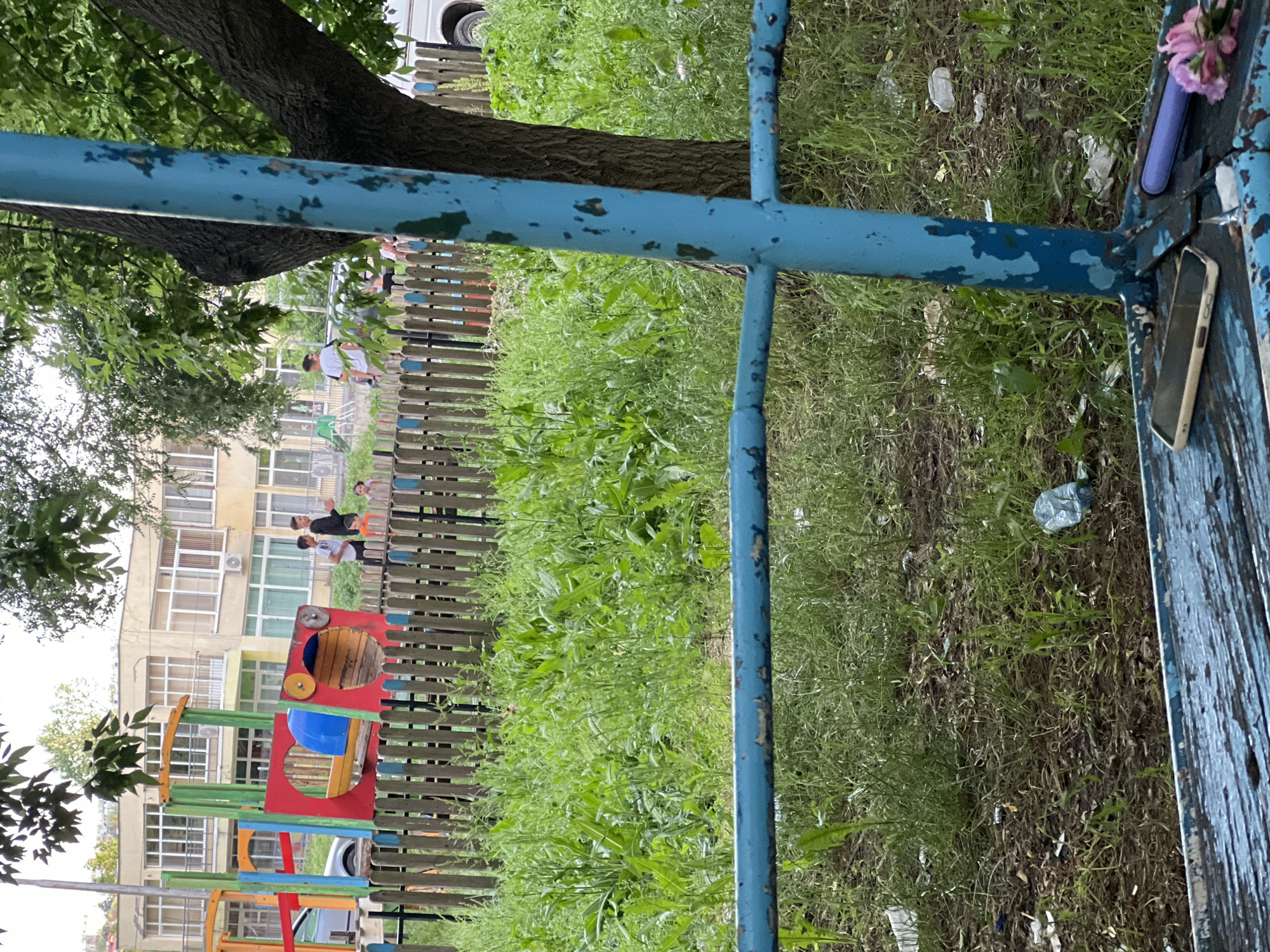 The image depicts an outdoor scene within the city of Dobrich, Bulgaria, featuring a playground area and residential or institutional buildings. In the immediate foreground, a weathered blue metal structure, possibly part of a swing set or bench, shows extensive paint chipping. Resting on its surface are a light-colored smartphone with a dark screen, a purple cylindrical object, and a small cluster of pink flowers. The ground directly beneath this structure is composed of dirt and patchy, overgrown grass, scattered with debris including plastic bottles and paper waste.
A wooden fence with alternating natural wood and faded blue slats runs horizontally across the midground, separating the foreground from an expanse of tall, unkempt green grass and weeds. Beyond this overgrown area, a colorful playground structure, predominantly red, blue, and yellow, resembling a train or multi-activity unit, is visible.
Several individuals are present behind the fence. From left to right: a person in a black hoodie, another in a light t-shirt, a small child in an orange top, another child in a light top, and two additional individuals in white t-shirts are seen standing or walking near the playground and fence line.
In the background, two multi-story buildings with light yellow facades, featuring numerous large windows and balconies, are visible. A large tree with green foliage obscures the right side of the scene, its dark trunk prominent. The foliage of another tree is visible in the upper left. A white van is partially visible on the far right, behind the tree and fence. The scene is illuminated by daylight, and the environment exhibits signs of disrepair, including chipped paint on structures and noticeable litter amidst the unmaintained vegetation.