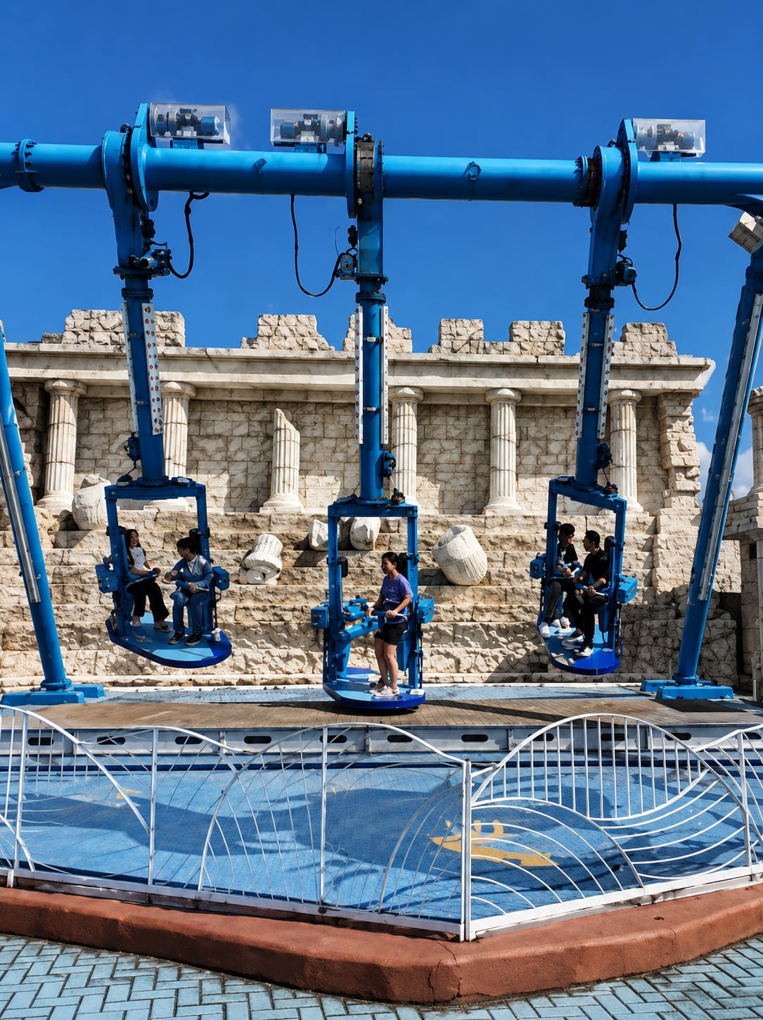 This image depicts an amusement park ride with a blue structure against a backdrop of a stone building with columns, resembling ancient ruins. The ride consists of several suspended seats, each carrying passengers, that swing in an arc. In the foreground, a blue platform with white fencing encloses a blue surface with yellow markings. The sky is clear and blue. This scene is located in the city of Berastagi, Indonesia.