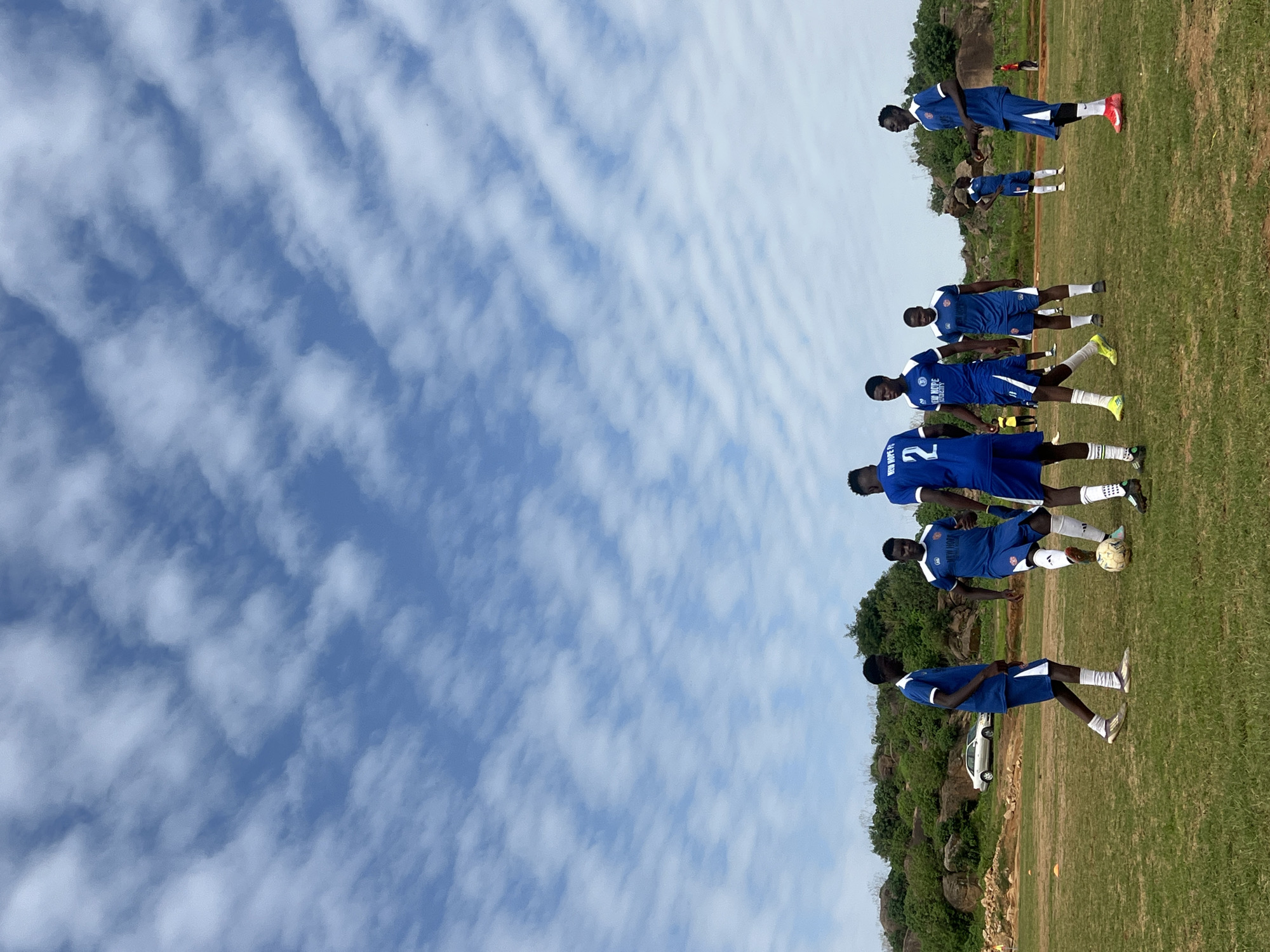 In Jos, Nigeria, a group of young men, dressed in matching blue and white football kits, engage in a practice session on a grassy field under a sky filled with scattered clouds. The players, varying in height and build, are positioned across the field, some in motion and others observing. One player in the foreground, wearing a jersey with the number 2, is the focus of attention. A soccer ball rests on the ground near two other players, one of whom appears to be in the process of kicking it. The background features a rugged, rocky hillside with sparse vegetation, and a silver sedan is parked on a dirt track adjacent to the field. The overall scene depicts a casual football training session in a somewhat undeveloped, outdoor setting.