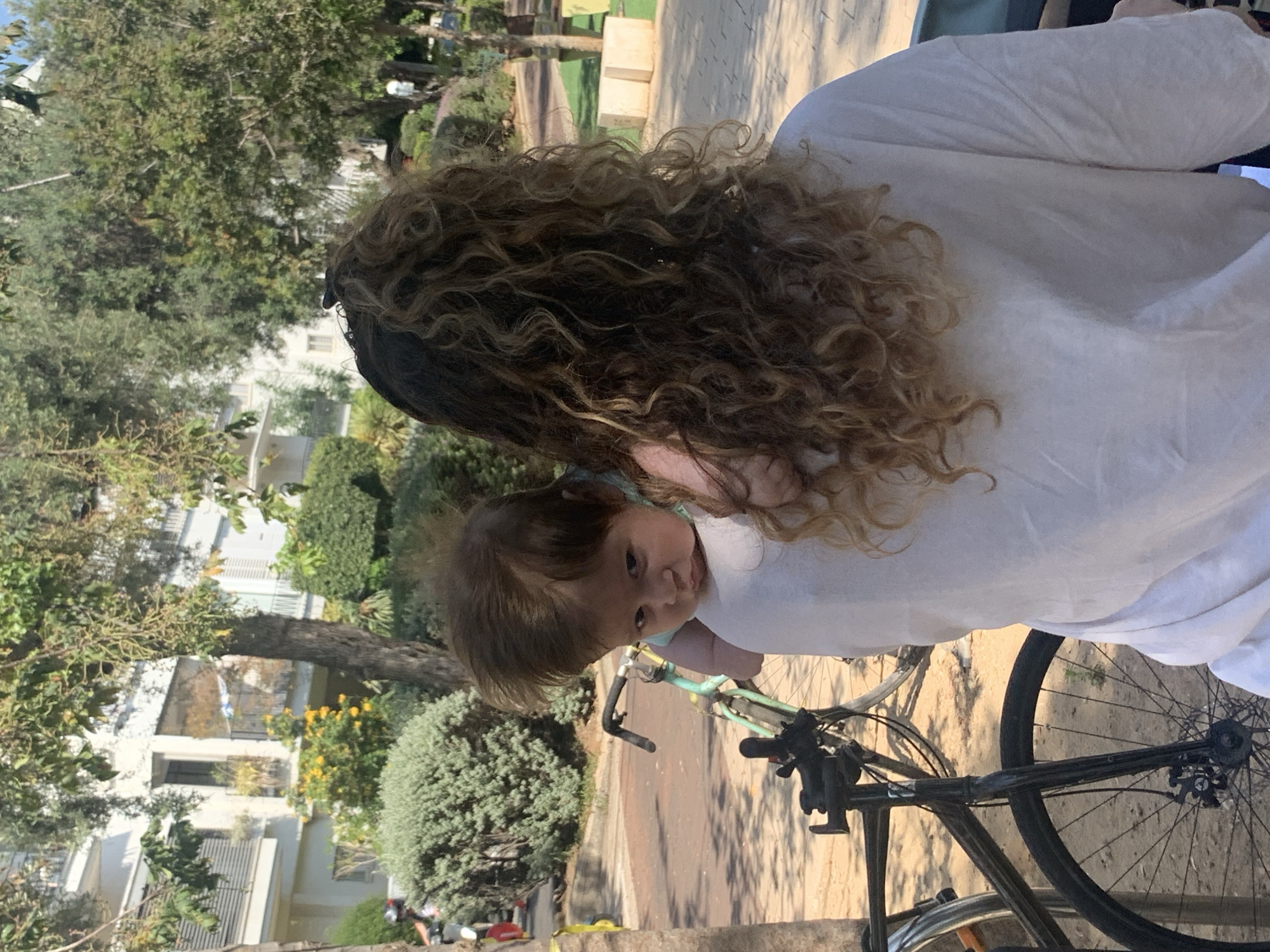 In Tel Aviv, Israel, a person with long, curly brown hair is carrying a baby on their left shoulder. The baby, facing away from the camera and looking to the side, has fair skin and messy brown hair. They are wearing a light-colored outfit. The person holding the baby is wearing a loose-fitting white t-shirt. In the background, there are trees with green leaves, a white building with balconies and windows, a light blue bicycle with a black handlebar, and another black bicycle leaning against a pole. The ground is paved with a light-colored surface.