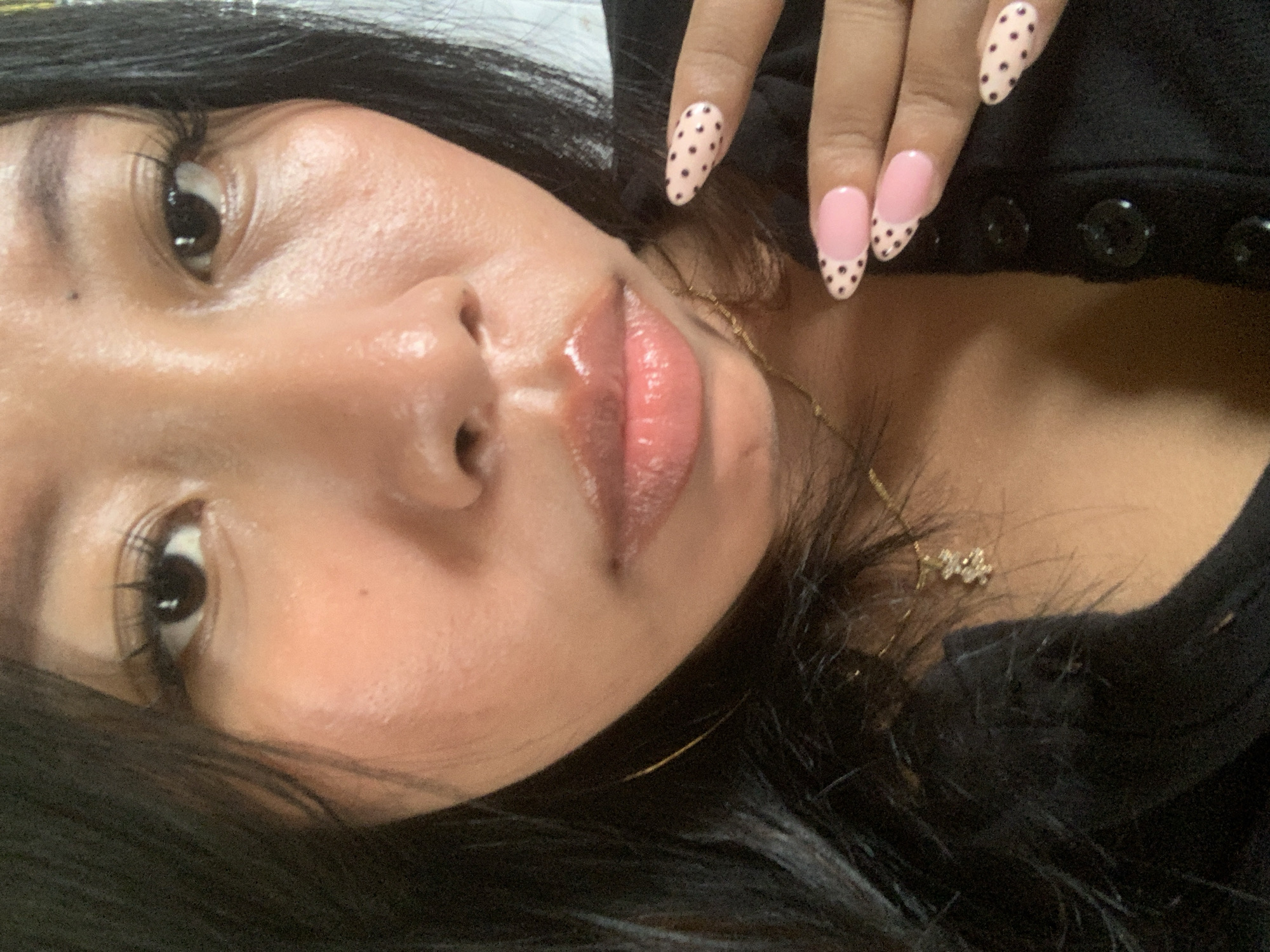 The image shows a close-up of a person's face and hands. The person has dark hair and is wearing makeup, including mascara and lip gloss. Their nails are painted pink with black polka dots. They are wearing a black shirt. The background is blurred. No specific location in Jajag, Indonesia, can be determined from the image.