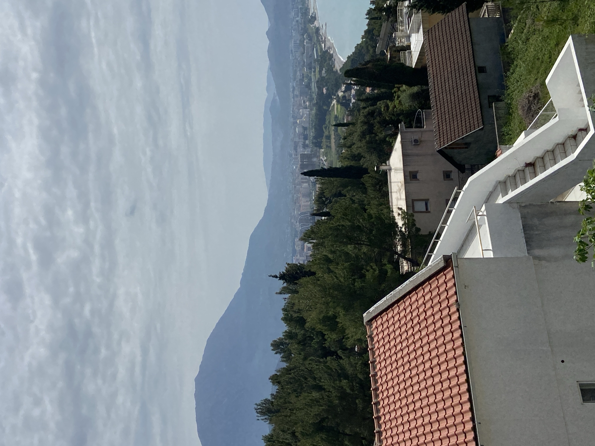 The image depicts a view from what is likely a residential area in Šušanj, Montenegro. In the foreground, there's a part of a building with a red tiled roof and a white exterior. A white staircase with railings is connected to the building. The middle ground is filled with dense green trees. Beyond the trees, the cityscape of Šušanj stretches towards the coastline. In the distance, there's a hazy mountain range under a cloudy sky, and a glimpse of the Adriatic Sea at the top right corner of the image.