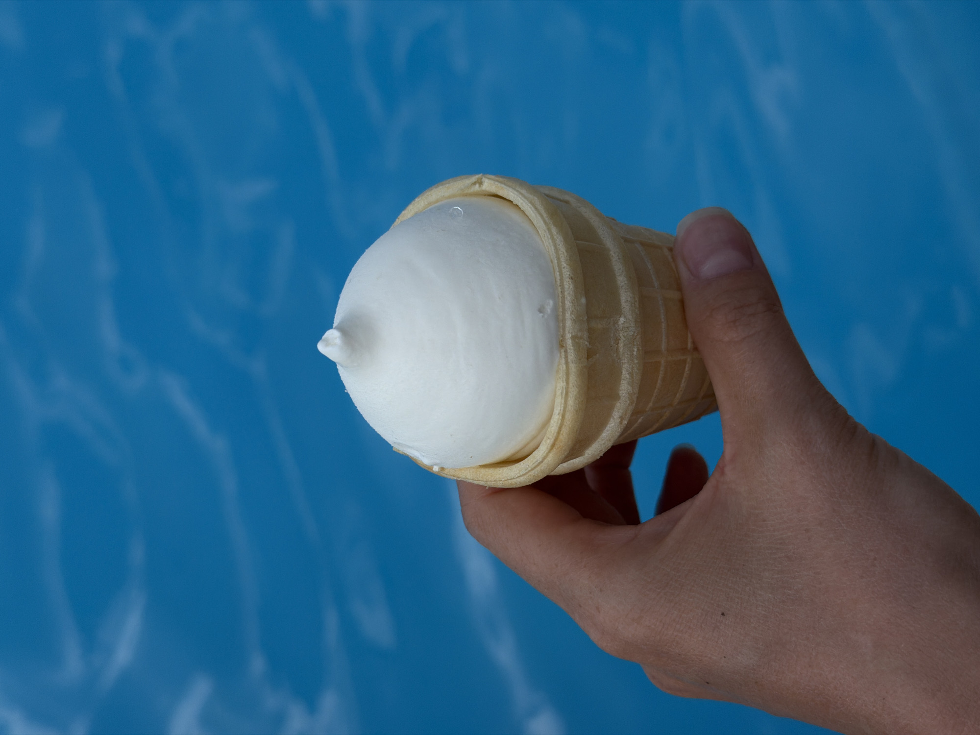 A hand is holding a vanilla soft-serve ice cream cone against a bright blue background. The ice cream has a smooth, rounded top with a slight peak. The waffle cone is light brown and has a grid pattern. The background appears to be water, possibly a swimming pool, with gentle ripples and highlights. There is no indication of the city of Kamyshin, Russia.