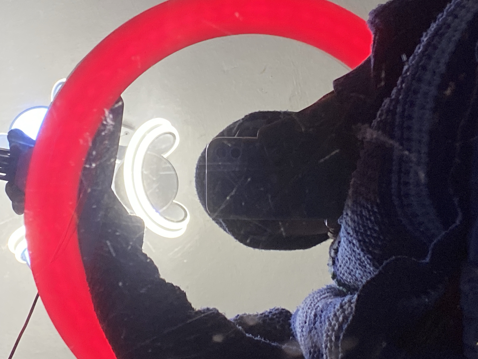 The image is a close-up, low-angle shot of a person's reflection in a mirror. The person is wearing a dark beanie and is holding a smartphone, also dark, in front of their face. The smartphone screen is reflecting the surrounding environment, showing the back of the phone with its camera lenses. The person is wrapped in a textured, dark blue knitted garment, possibly a blanket or sweater. In the background, a bright red, circular light fixture, likely a ring light, is prominently featured. Above and behind the person's head, a white, curved fluorescent light is visible, suggesting the environment is indoors, possibly a room with artificial lighting. The overall impression is of a person taking a selfie or recording a video, illuminated by a ring light. The context of Abakaliki, Nigeria, is not evident from the visual information in the image.